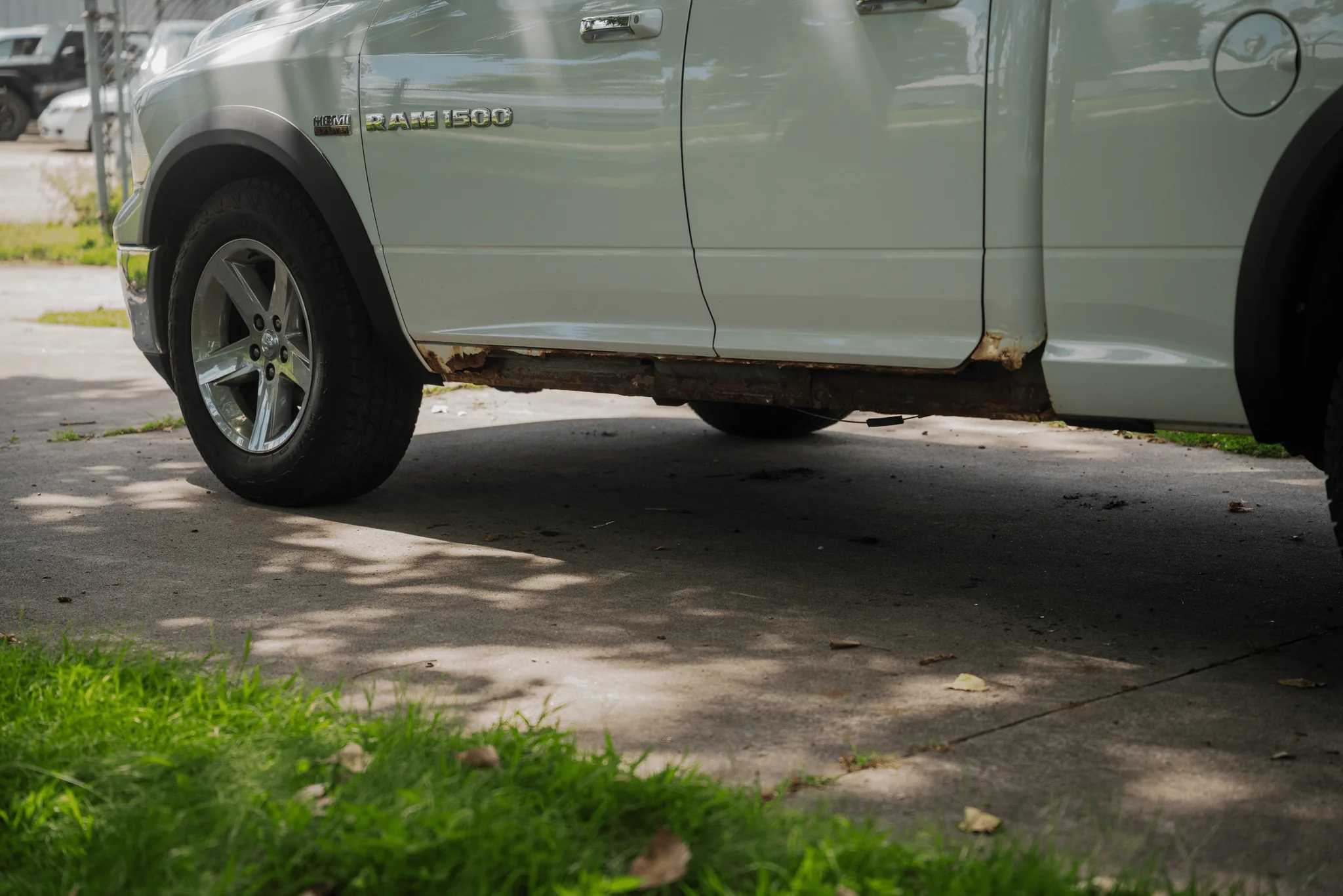 A silver Ram 1500 pickup truck with visible rust and damage beneath the driver's side door, parked on a concrete surface.
