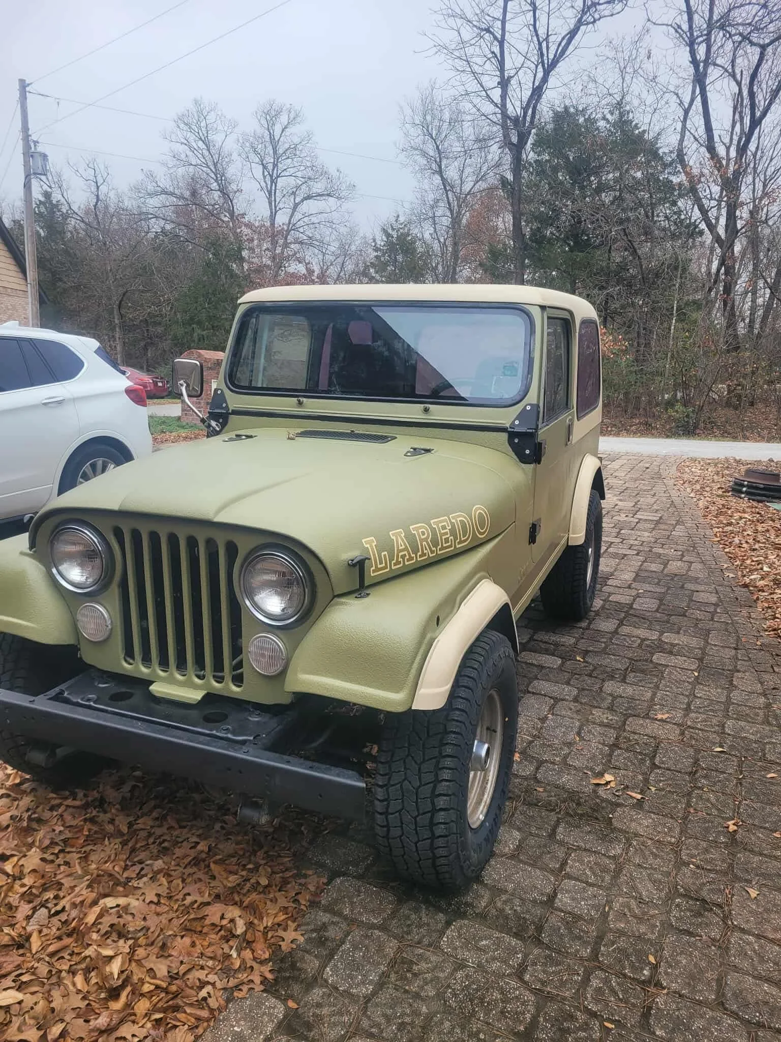A vintage olive green Jeep with the word 'LAREDO' written on the side, parked on a brick driveway surrounded by fallen autumn leaves, with trees and other vehicles in the background.