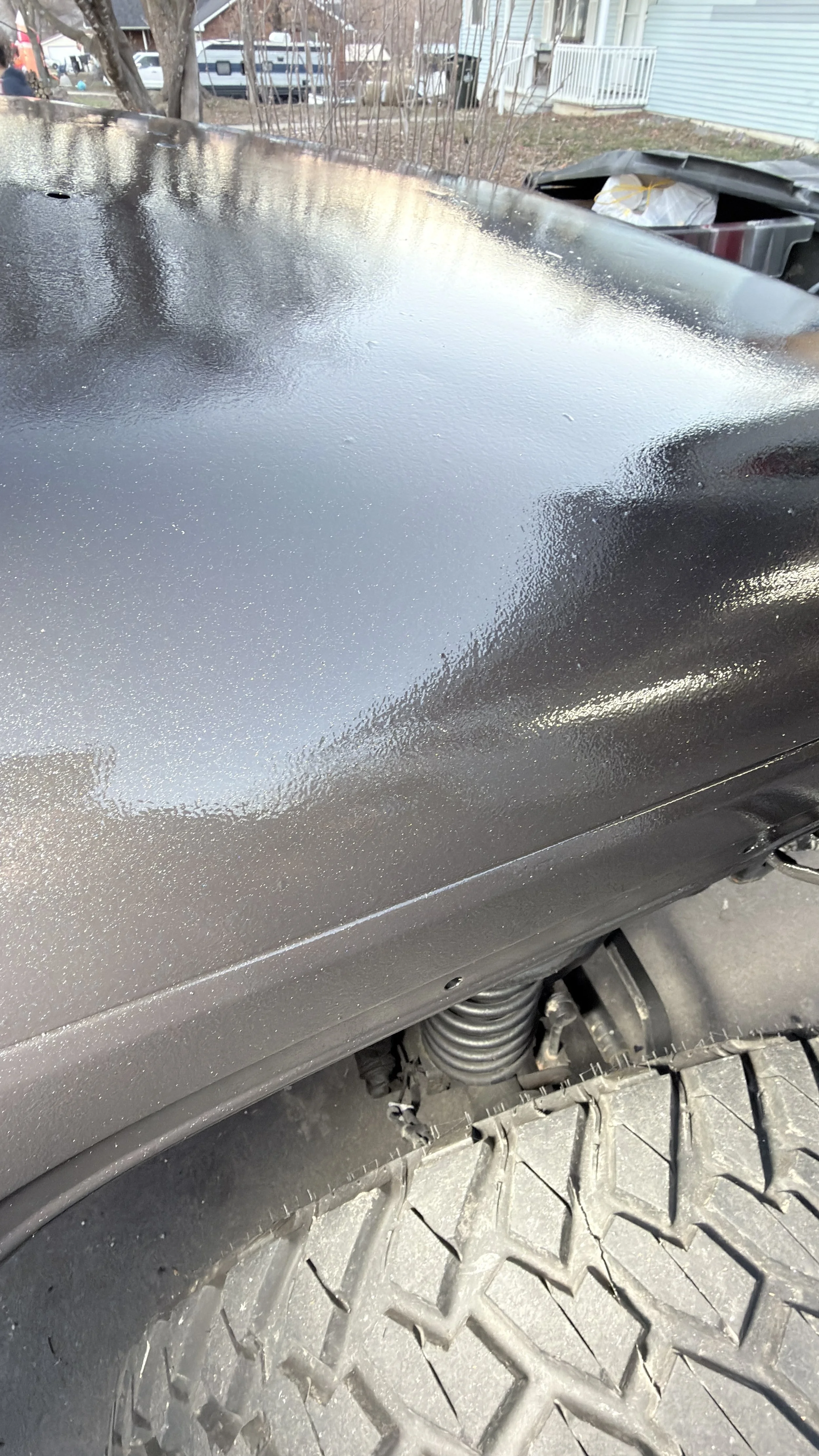 Close-up of a vehicle's black painted and textured fender and tire, parked outdoors in a residential area with trees, houses, and garbage bins in the background.