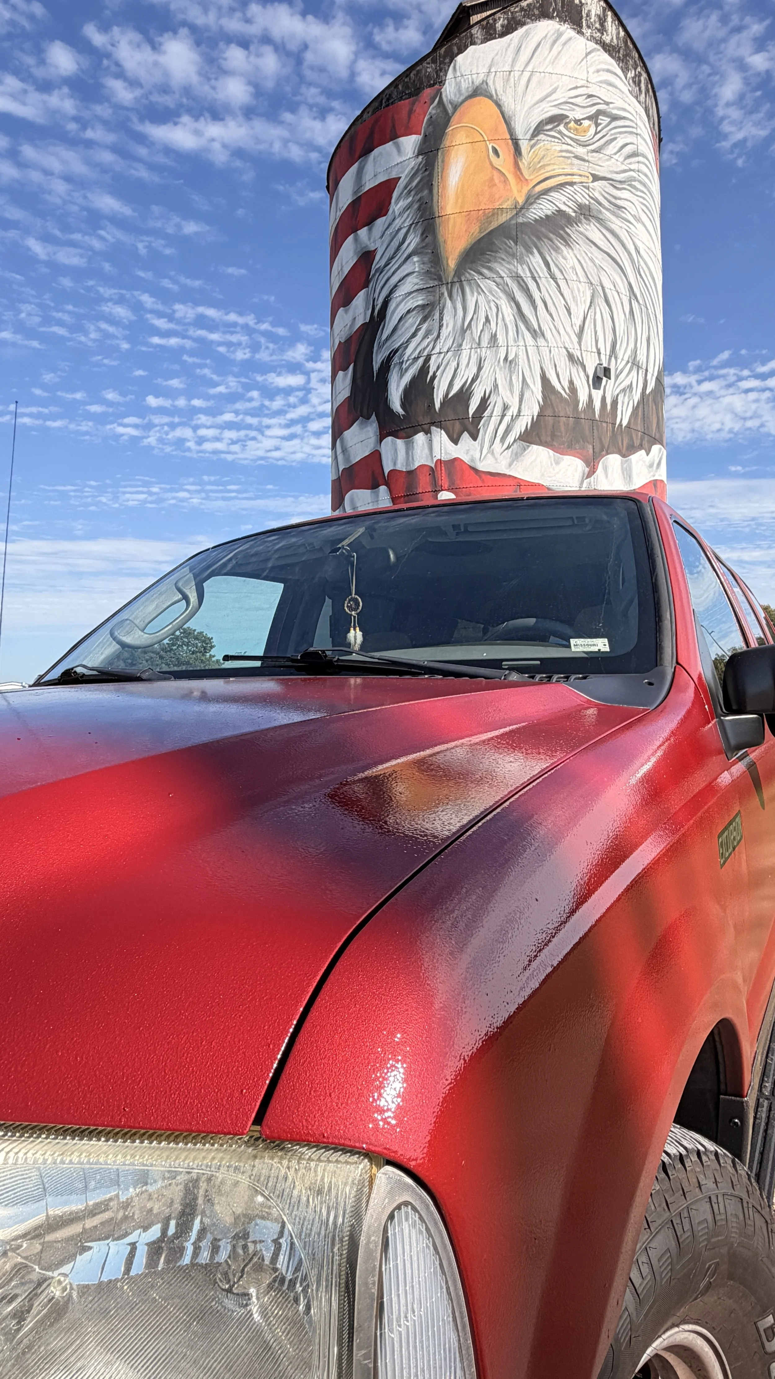 A red pickup truck parked outdoors with a large bird mural painted on a cylindrical structure behind it. The mural depicts an eagle wearing a red, white, and blue headband and the background features a partly cloudy blue sky.