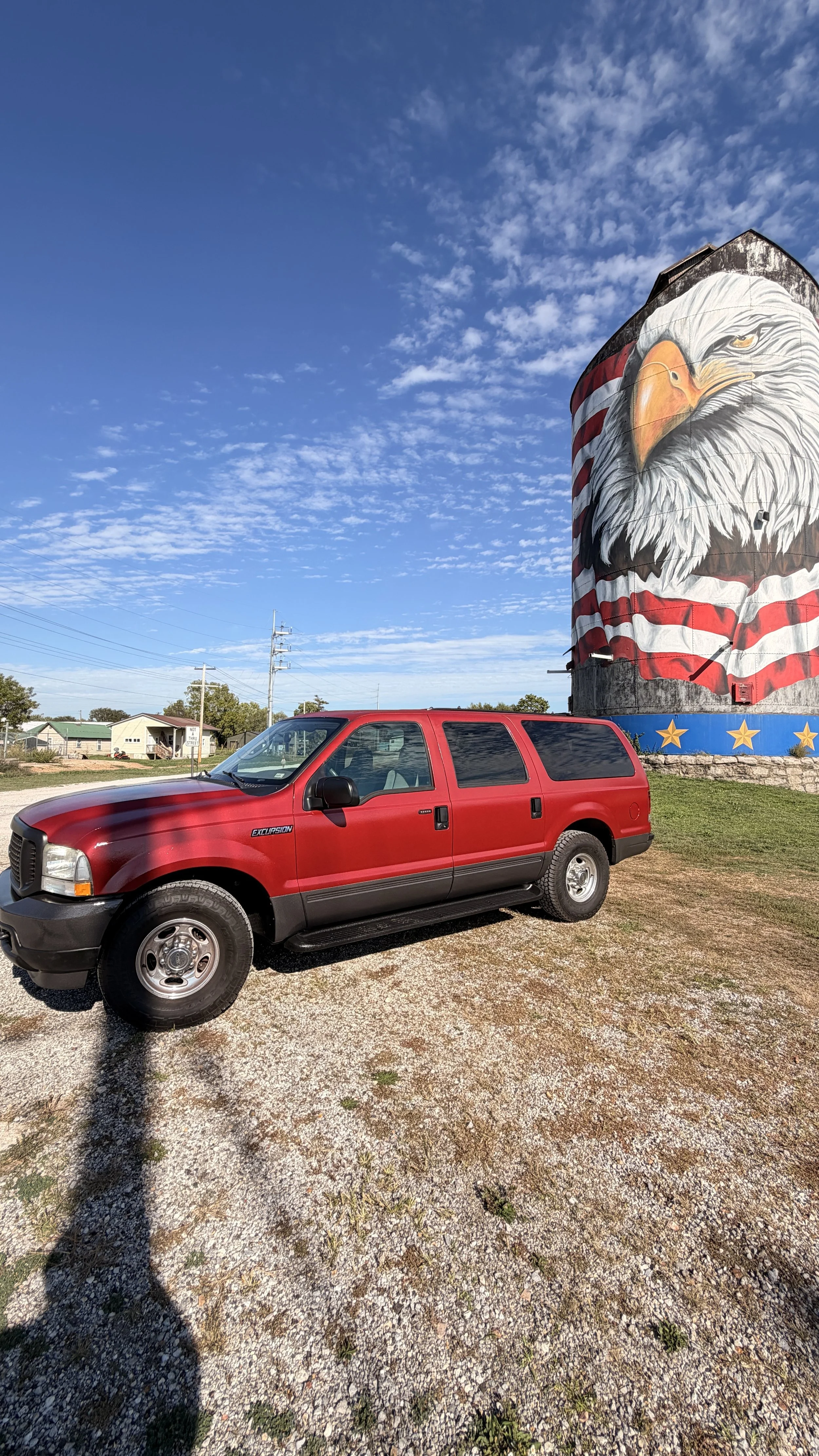 A red SUV parked on a gravel lot with a large mural of an American eagle painted on a silo in the background. The sky is blue with scattered clouds.