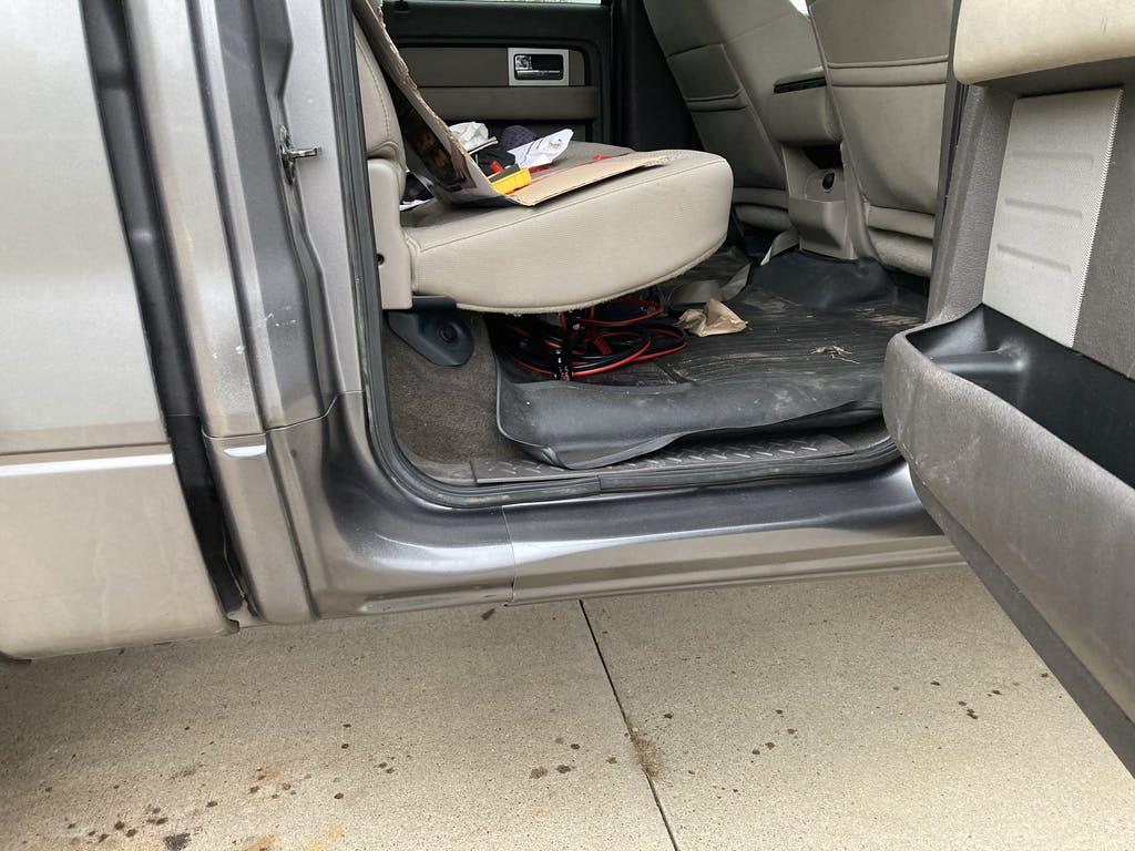 The interior of a vehicle showing the front passenger seat area with the door open. There are tools and items on the seat, and wiring visible underneath the seat. The ground outside the vehicle is concrete.