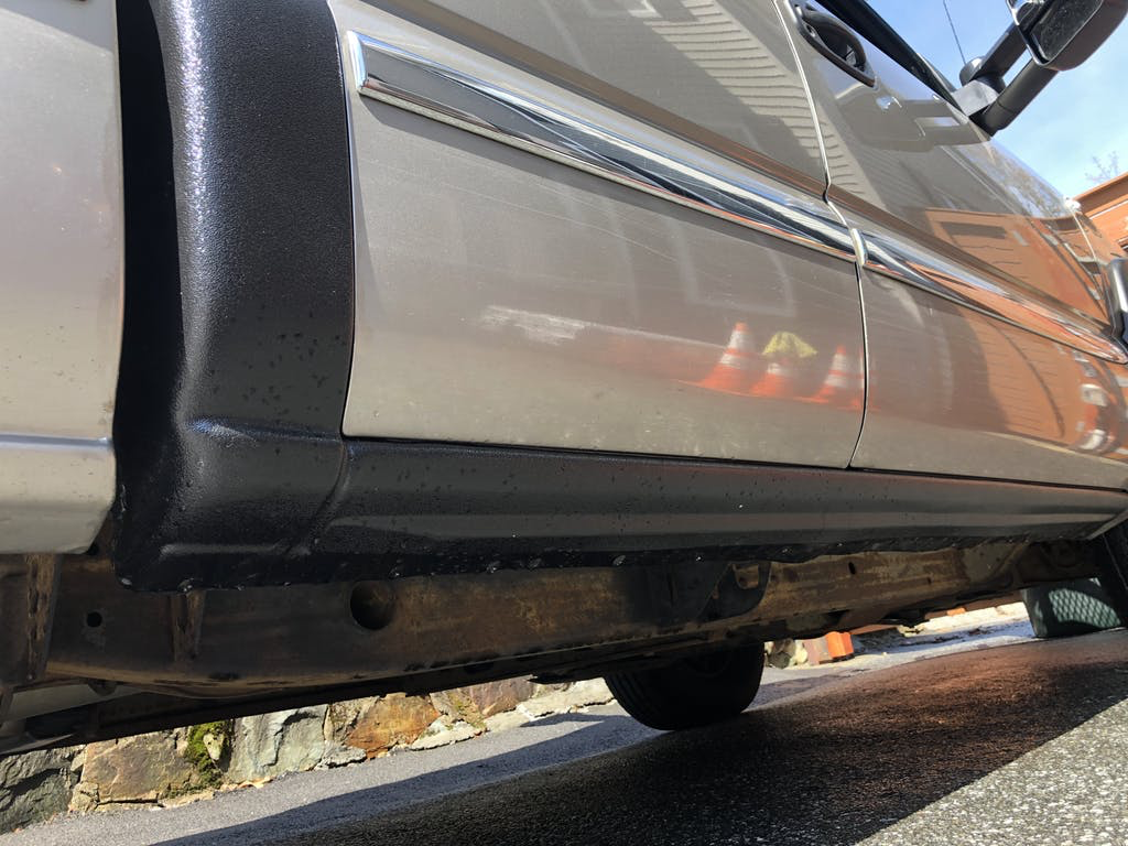 Close-up of the lower side of a vintage silver car showing the black running board and part of the tire, with a stone wall in the background.