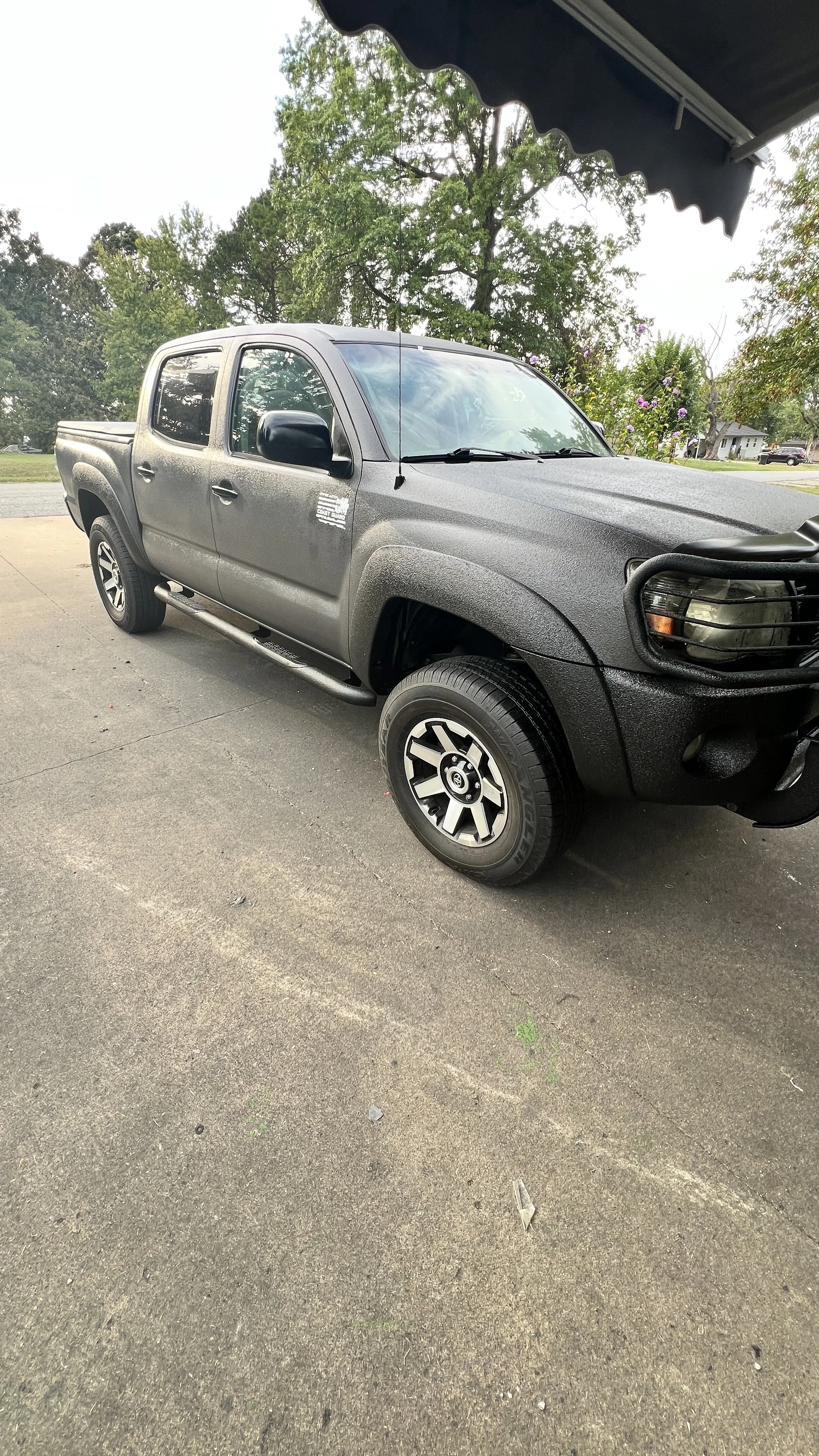 A dark gray pickup truck parked on pavement under a canopy, with trees and a house in the background.