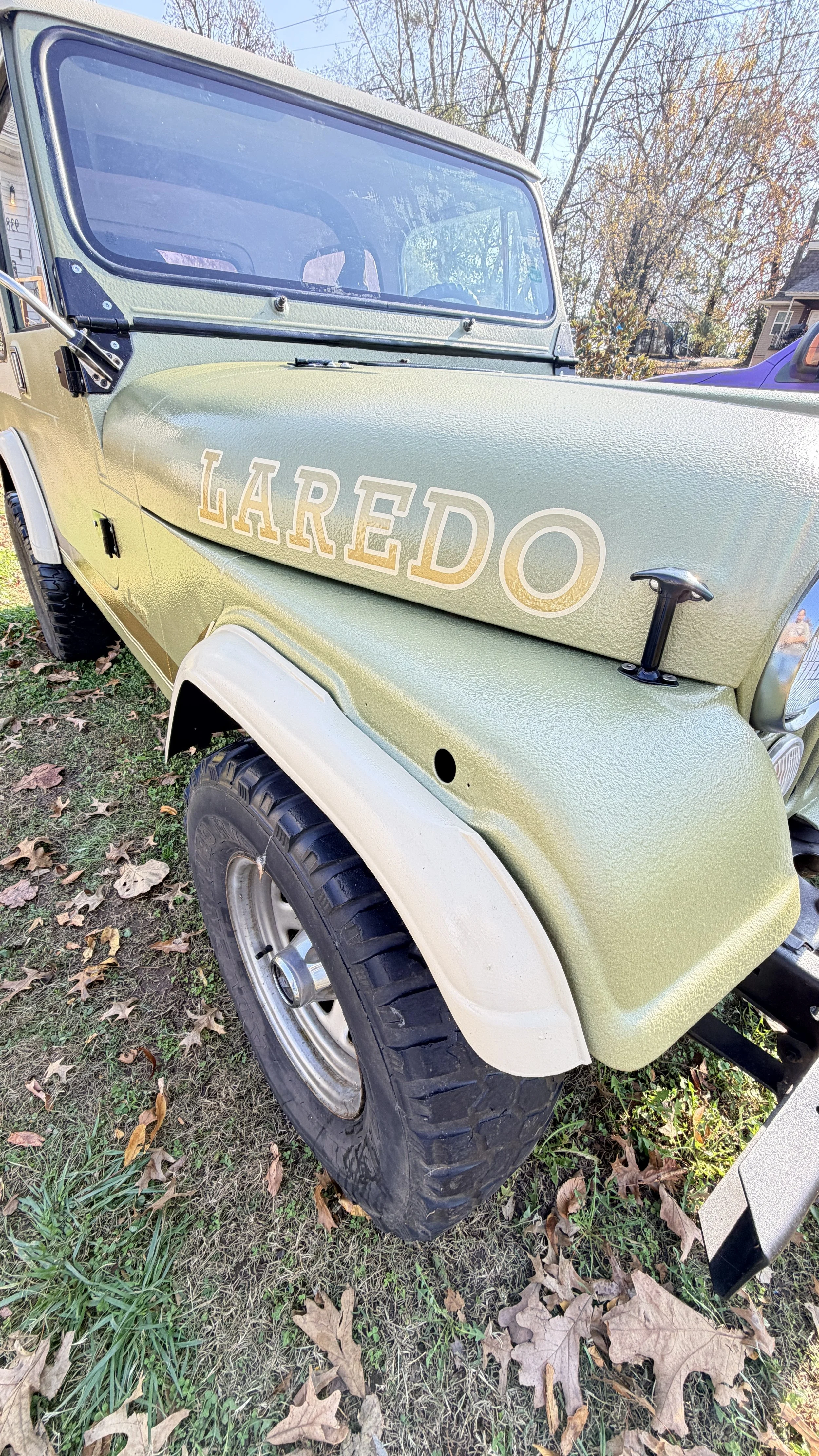 Close-up of a green off-road vehicle with the word 'LAREDO' on the side, parked on grass with fall leaves, trees, and houses in the background.