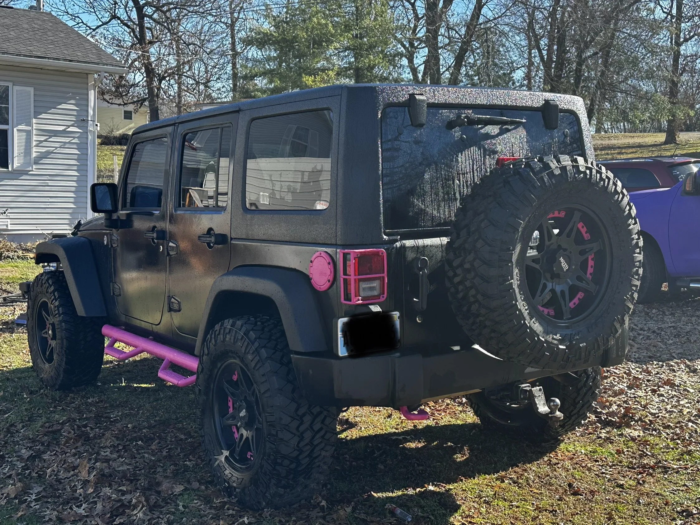 Black off-road Jeep with purple accents parked on grass near a house, with trees in the background.