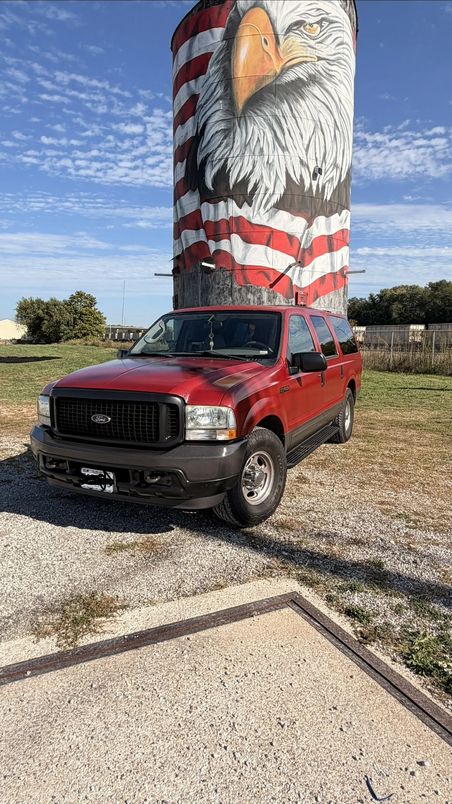 A red Ford SUV parked outdoors in front of a tall, cylindrical water tower painted with a mural of a bald eagle's face, with a blue sky and some clouds in the background.