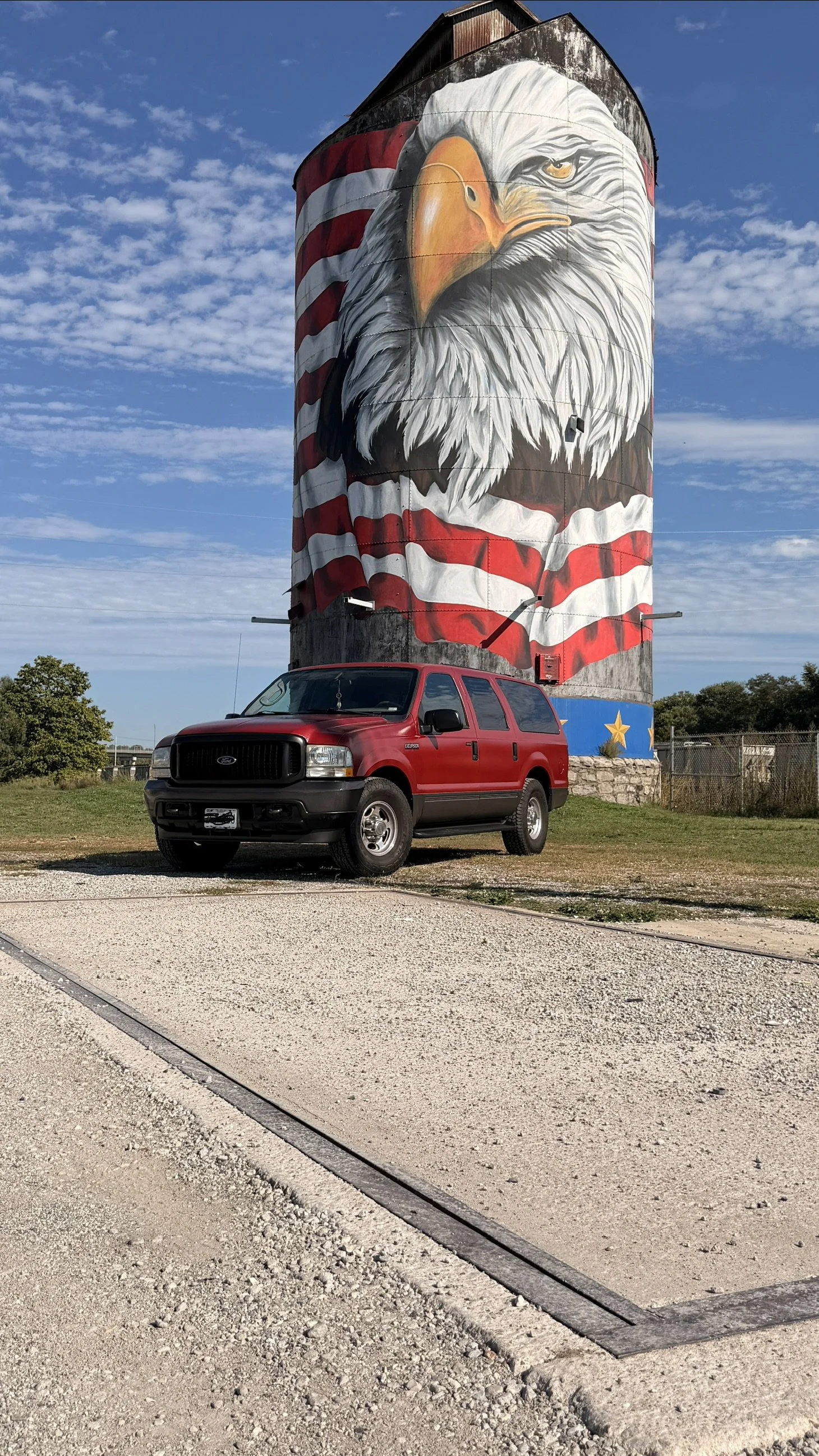 A red SUV parked on a gravel area in front of a large silos with a mural of a bald eagle wrapped in an American flag.