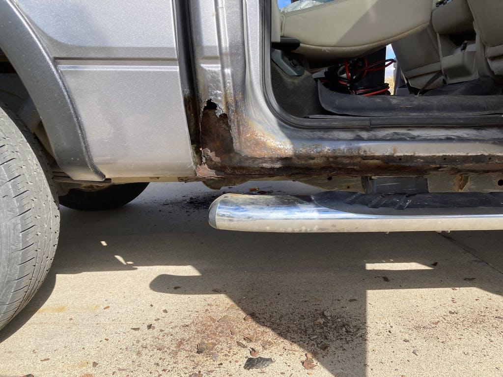 The image shows the lower side of a vehicle with rust and damage near the door frame. There is a chrome side step underneath and a partial view of a tire on the left. The interior of the vehicle is visible through the open door, and there is some dir