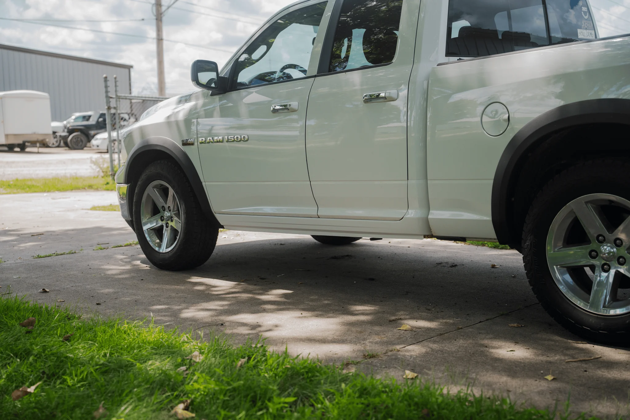 Side view of a white RAM 1500 pickup truck parked on a concrete surface with green grass and trees nearby, and other vehicles and structures in the background.