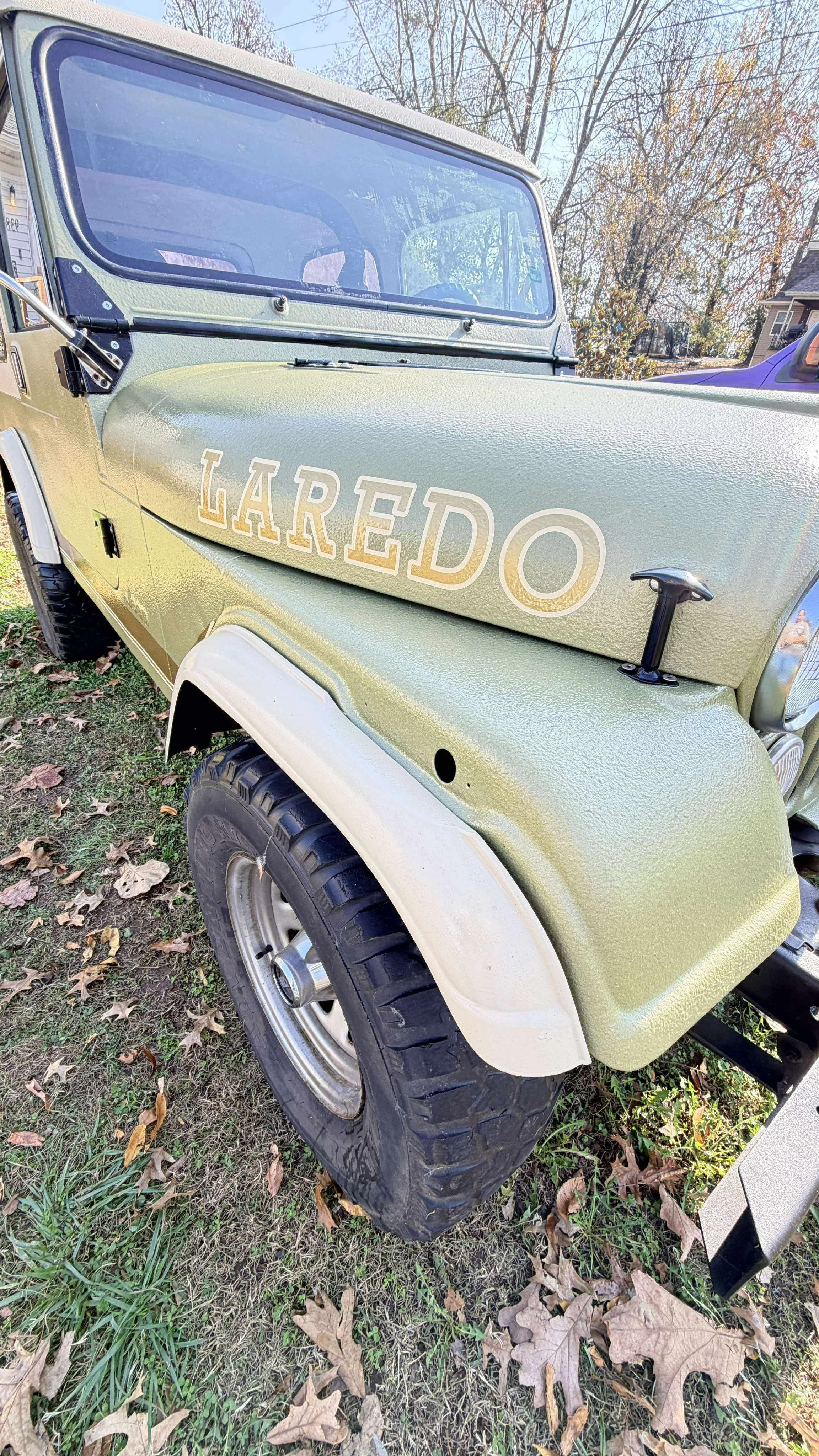 Close-up of a vintage green off-road vehicle with the word 'LAREDO' on the front side panel, parked on grass with fallen leaves.