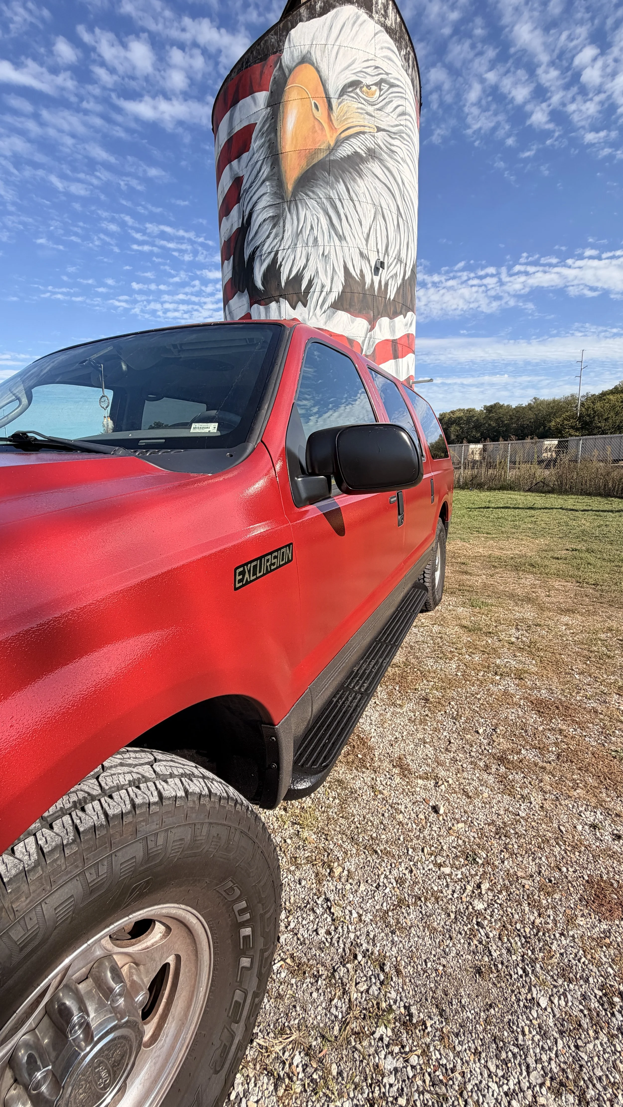 A red Ford Excursion vehicle parked in front of a large painted silo with an American flag-themed eagle mural.