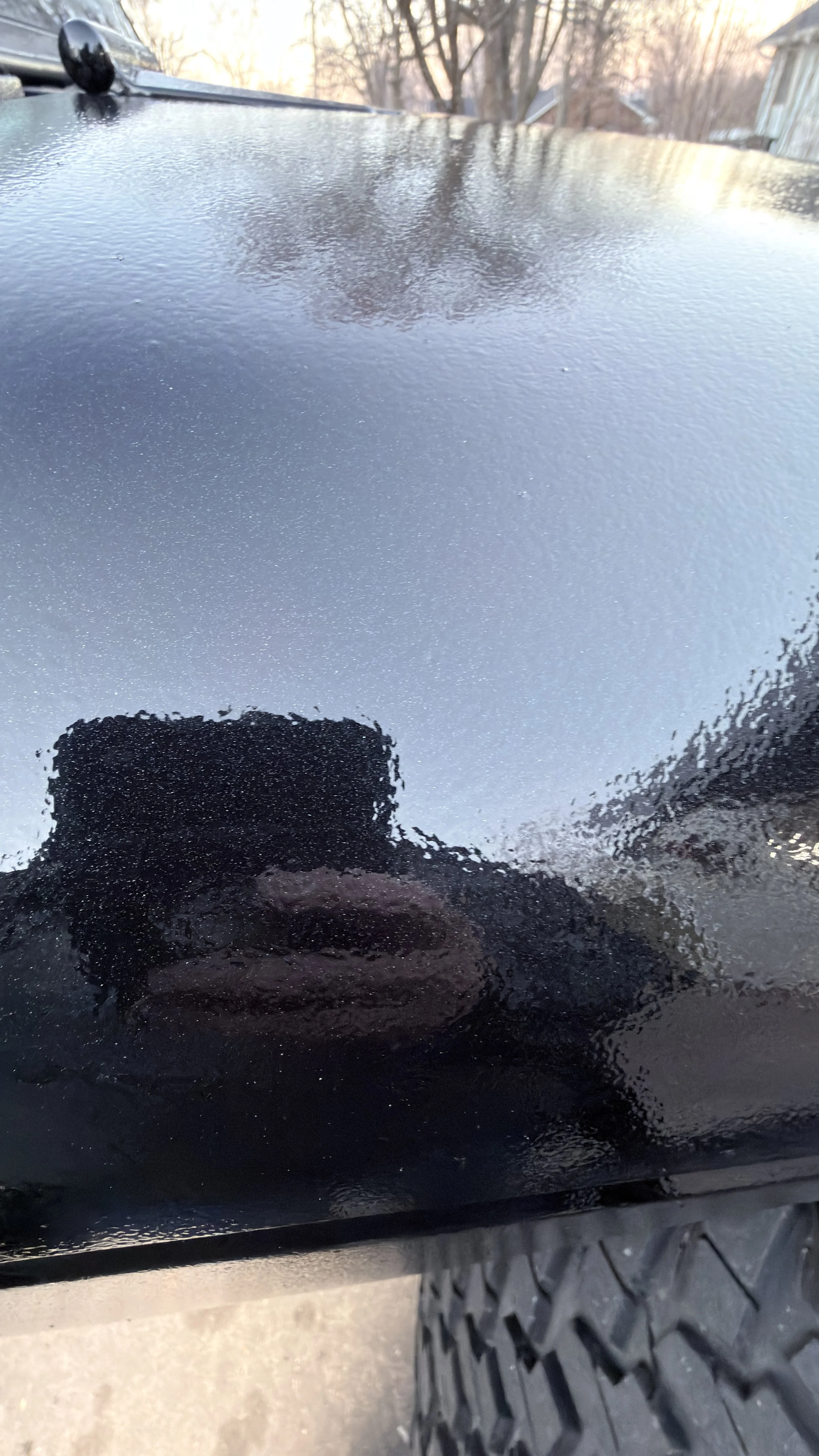 Close-up of a black, textured, and slightly reflective surface, likely part of a vehicle, with a reflection of the person taking the photo and an outdoor background with trees and houses.