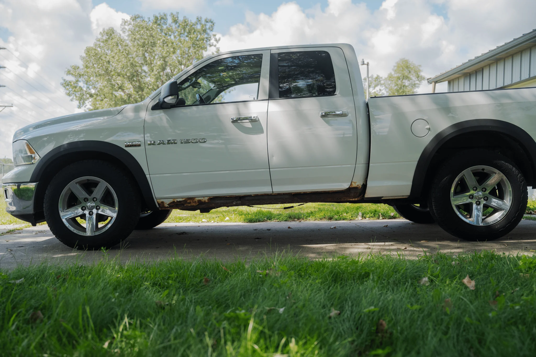 Side view of a white pickup truck with visible rust and damage beneath the doors, parked on a driveway with grass in the foreground and a house to the right. Trees and cloudy sky in the background.