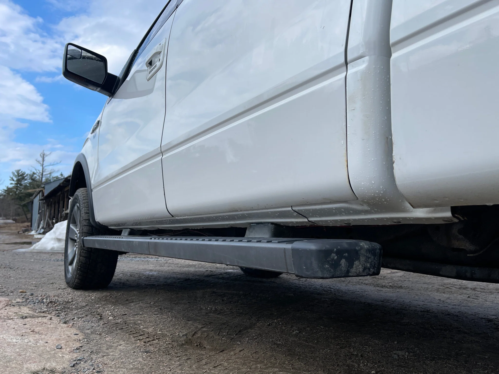 Close-up of the side of a white pickup truck, showing the running board and part of the tire, with a dirt ground and a blue sky with clouds in the background.