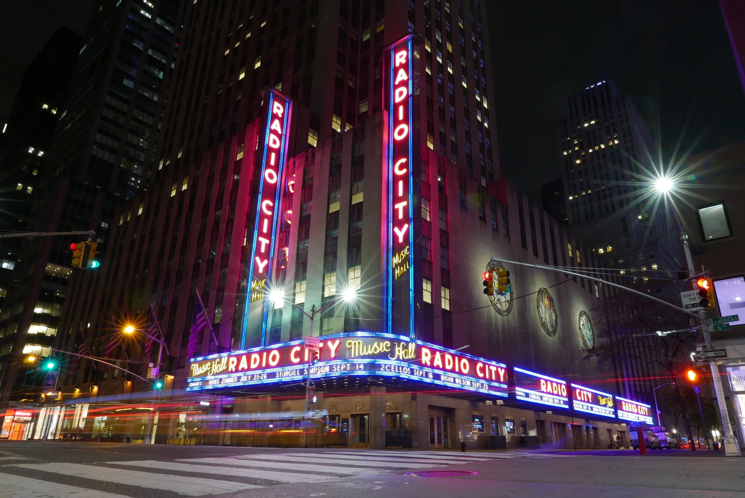 Nighttime view of the Radio City Music Hall in New York City with bright neon lights and billboards on the building facade.