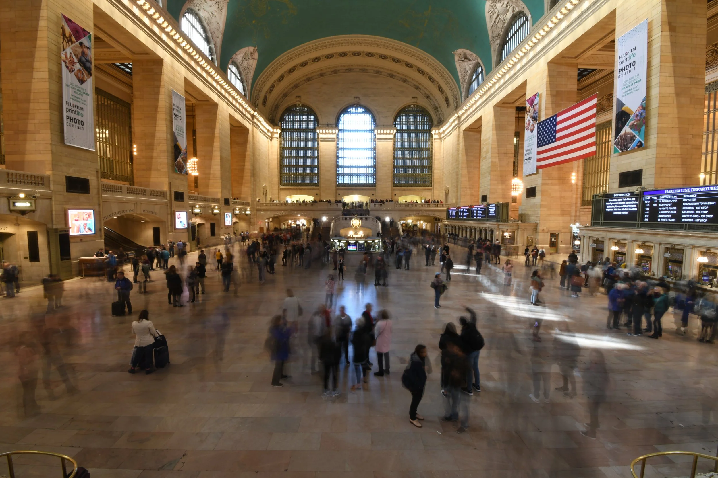 Interior of Grand Central Terminal's main hall with people walking and standing, large windows, high arched ceiling, American flag, electronic departure boards, and banners.
