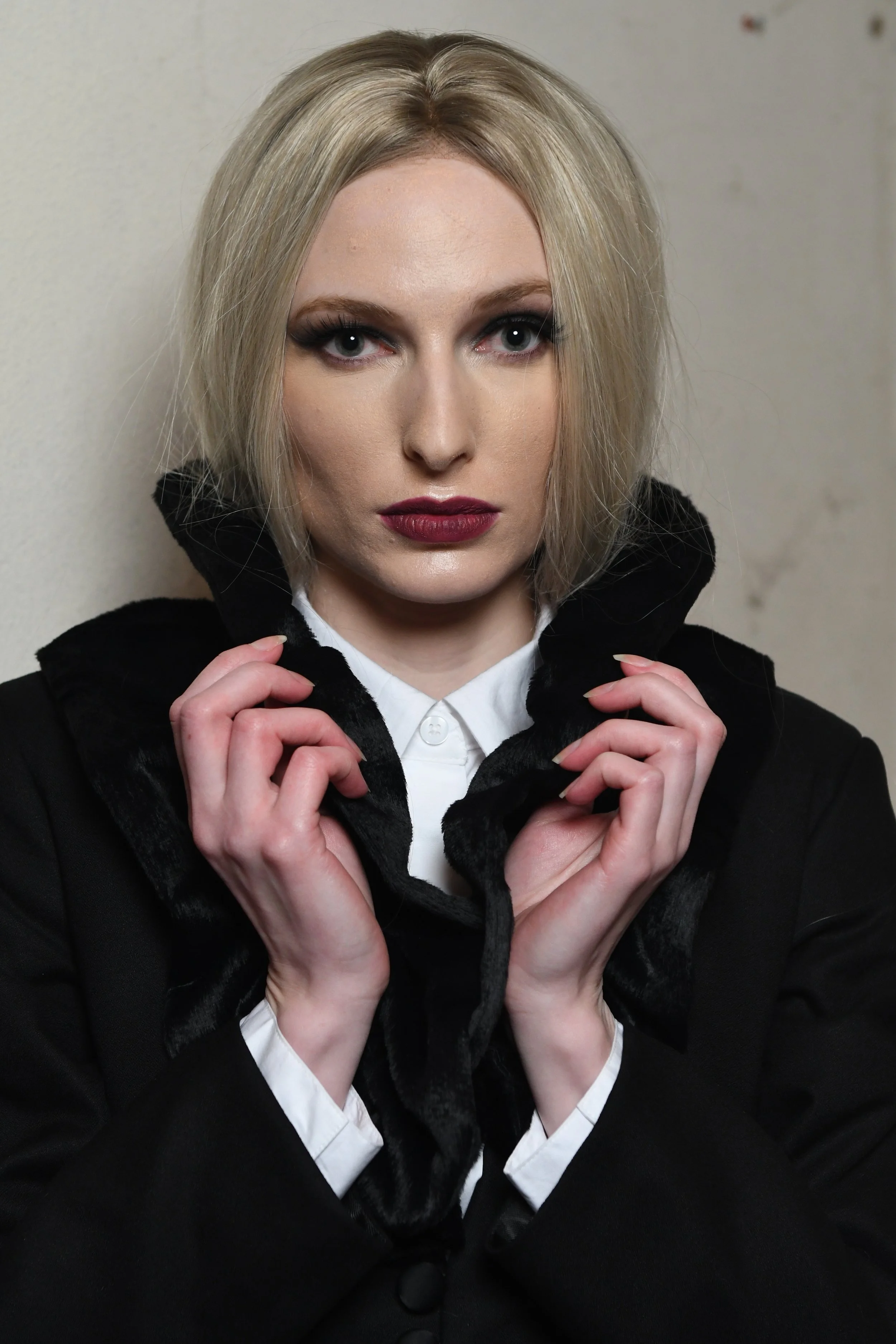 A woman with platinum blonde hair and dark makeup holding her collar and looking directly at the camera.