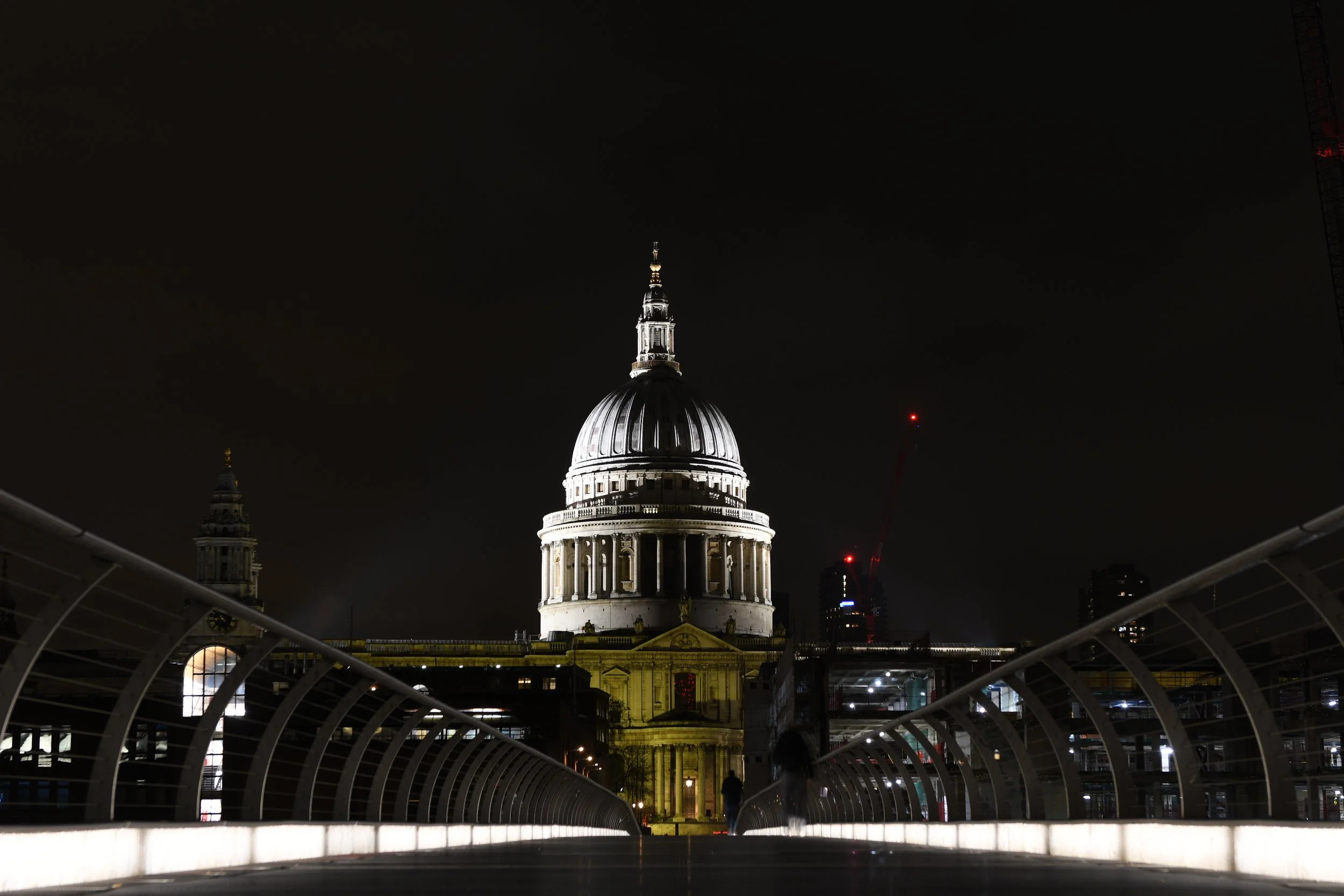 Night view of St. Paul's Cathedral illuminated from below, seen from a pedestrian bridge with glowing walkway railings.