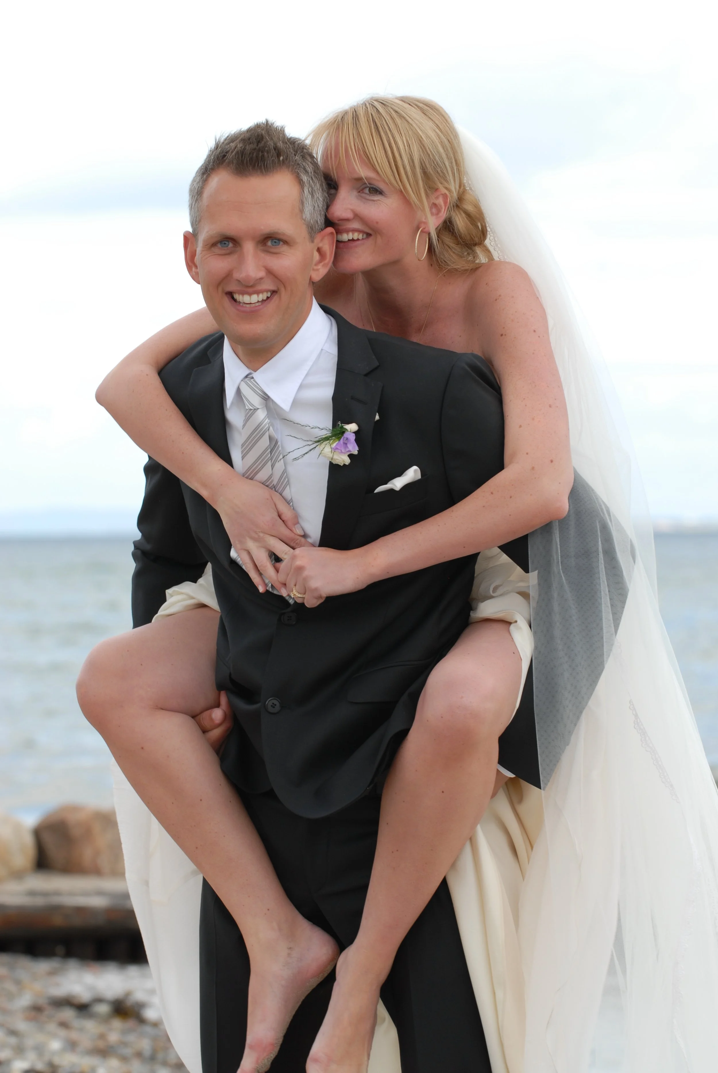 A newlywed couple at the beach, with the groom carrying the bride on his back and both smiling.