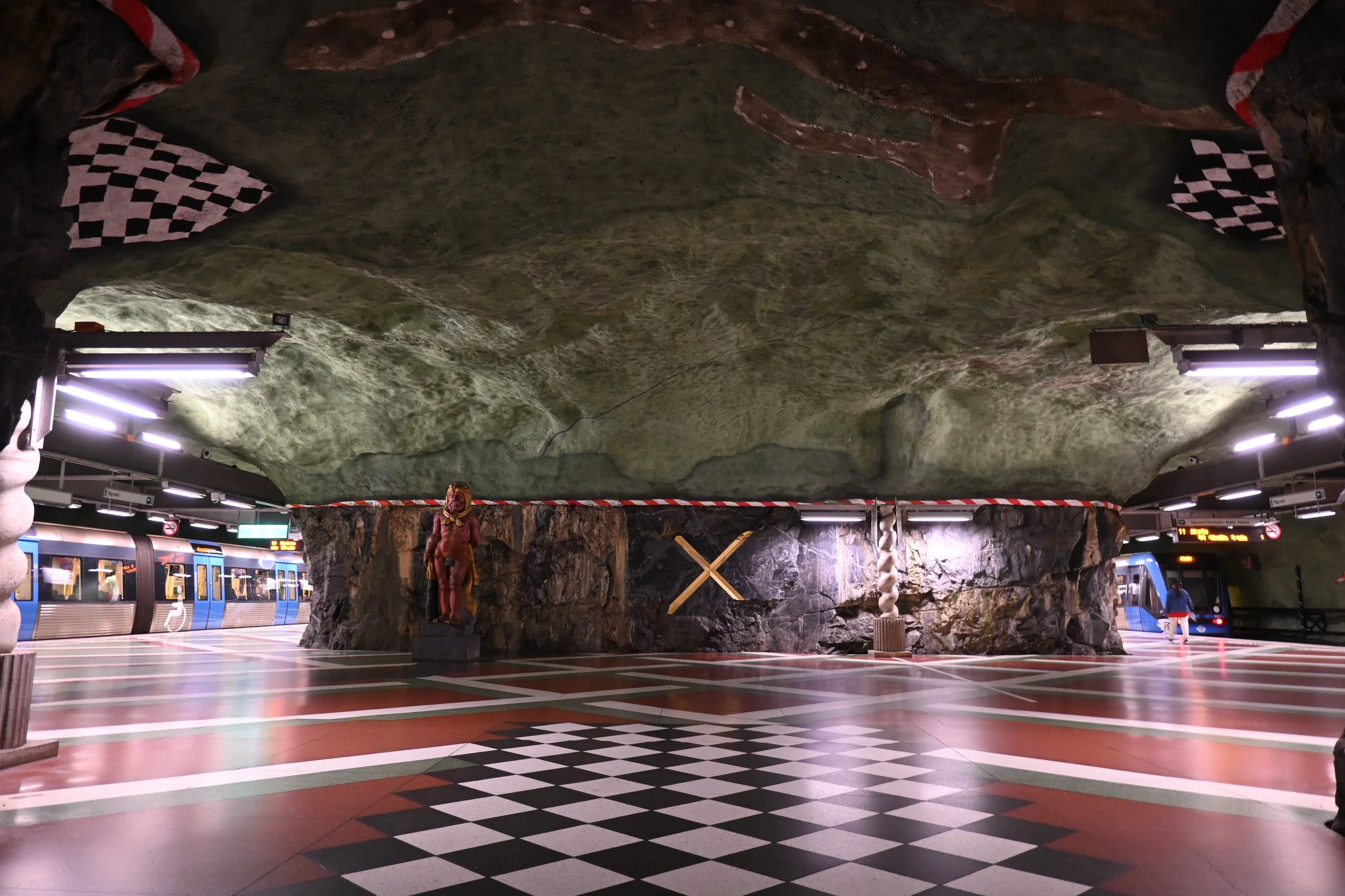 Underground subway station with a large artificial cave ceiling, checkered pattern floor, statue of a lion with a lion's head on a pedestal, and a train arriving on the platform.