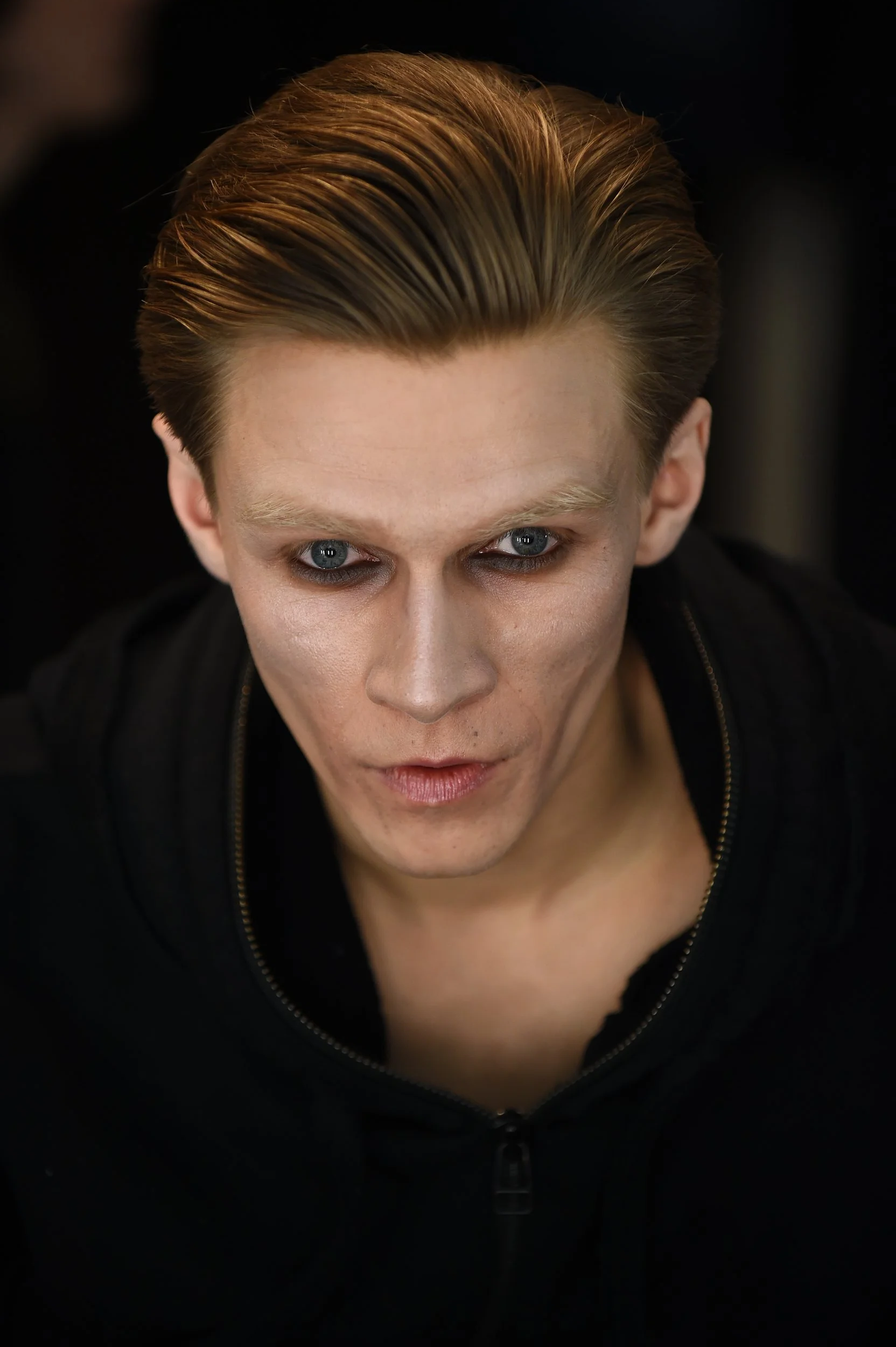 Close-up of a man with slicked-back blond hair and blue eyes, wearing a black shirt with an extended collar.