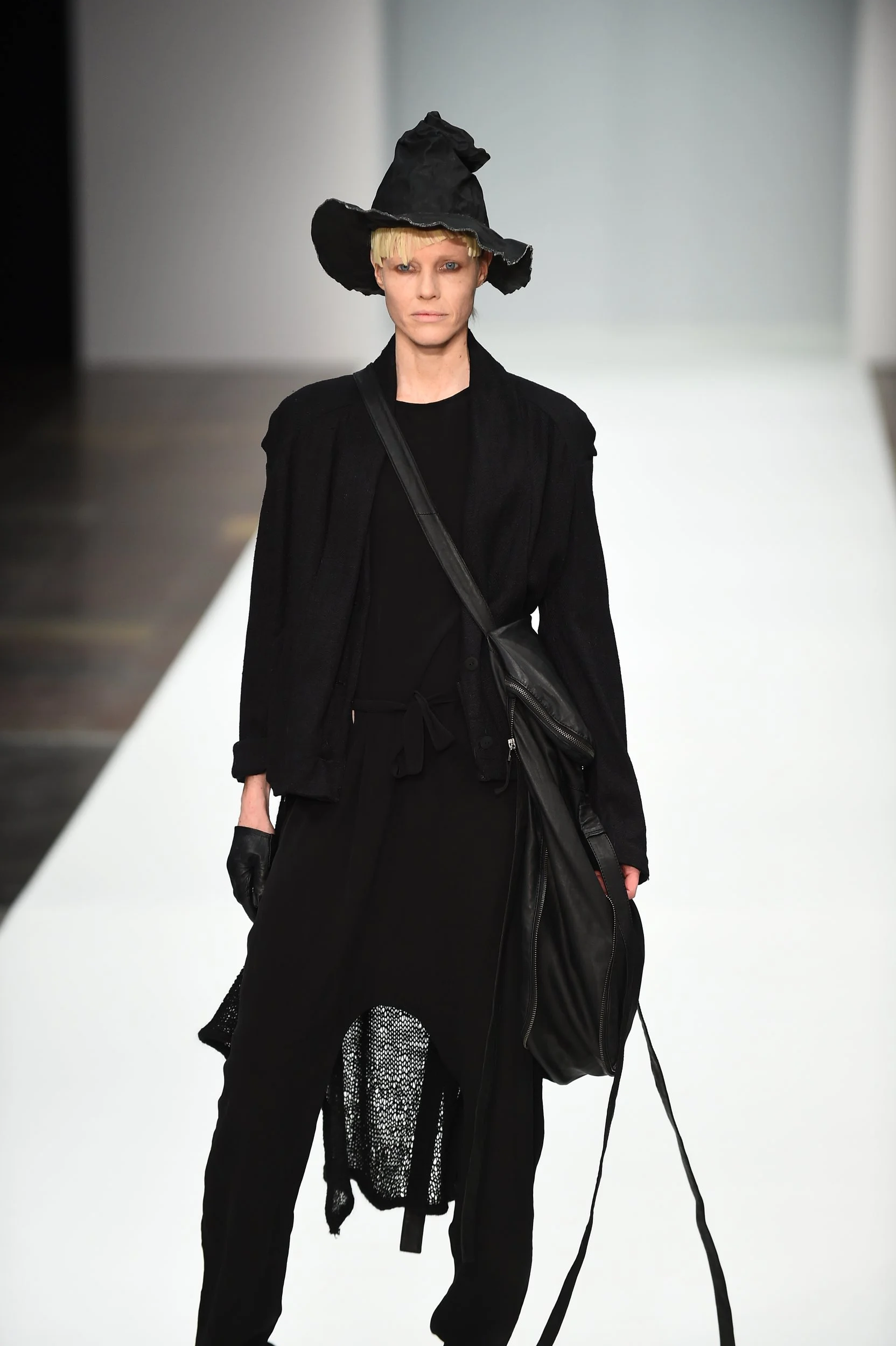 Model walking down a runway dressed in black clothing, wearing a large, floppy black hat and carrying a black bag.