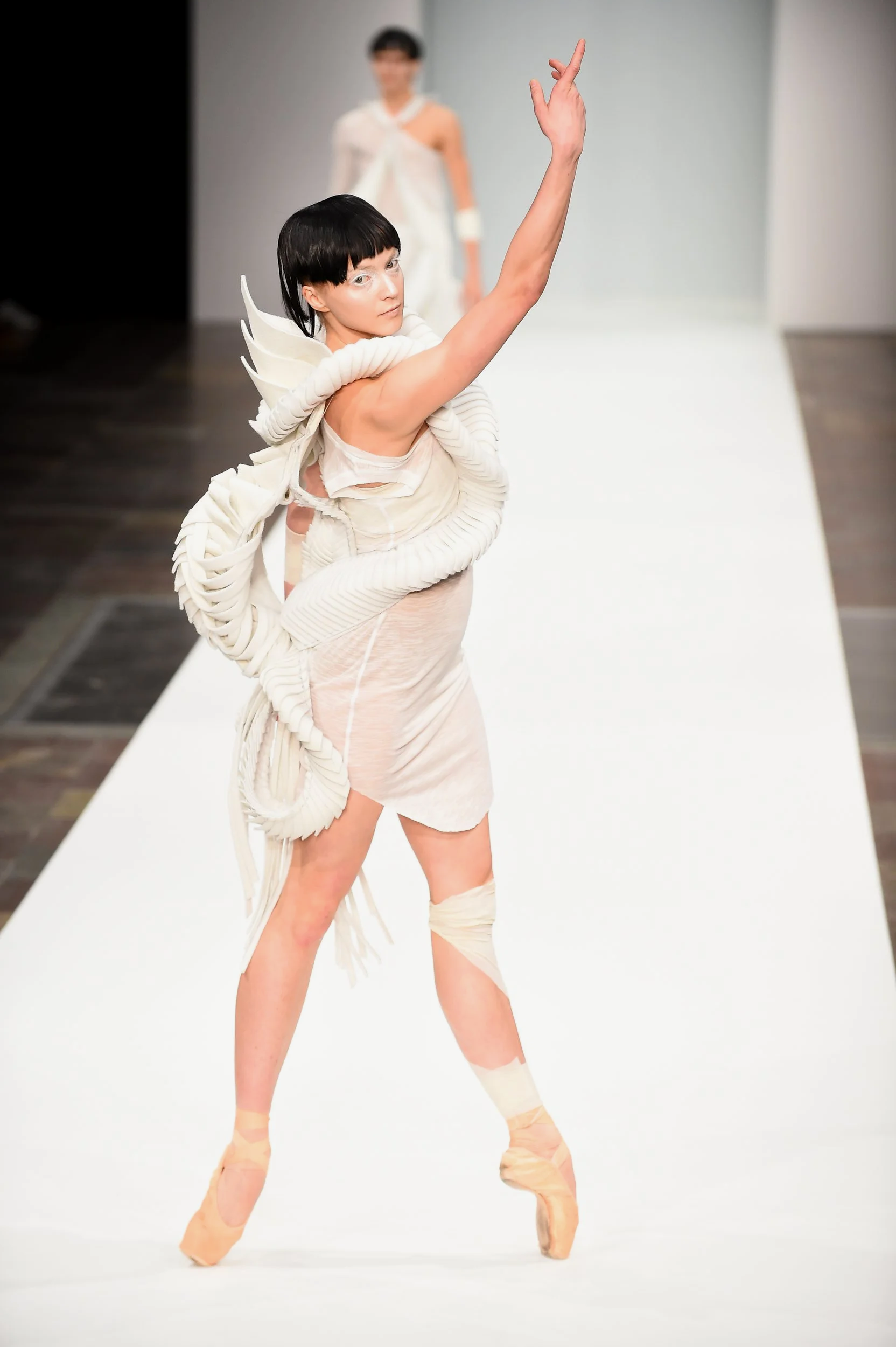 Model walking on runway in avant-garde white costume with sculptural details, wearing ballet shoes, with another model blurred in the background.