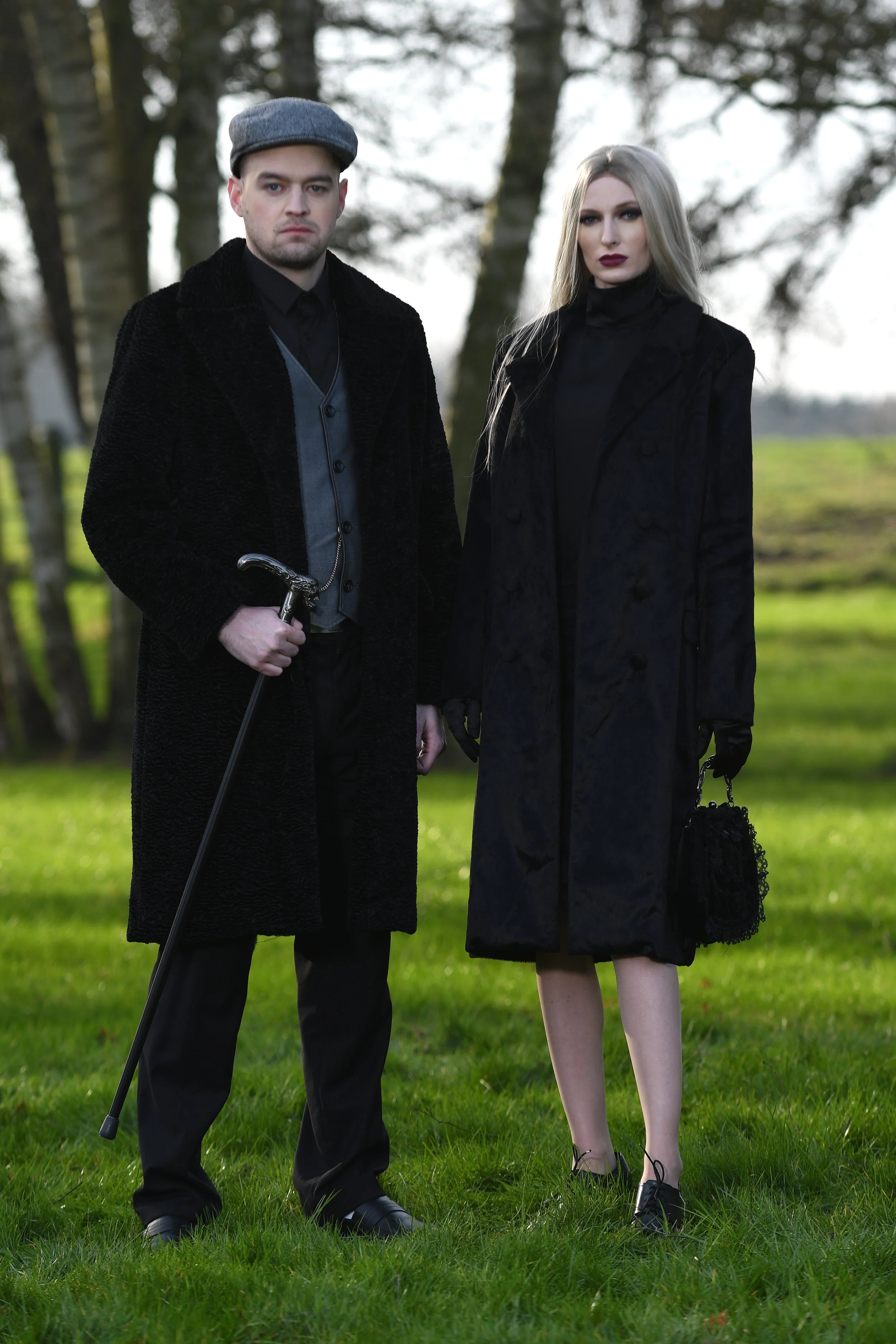 Man and woman dressed in dark, vintage clothing standing outdoors on grass with trees in the background.