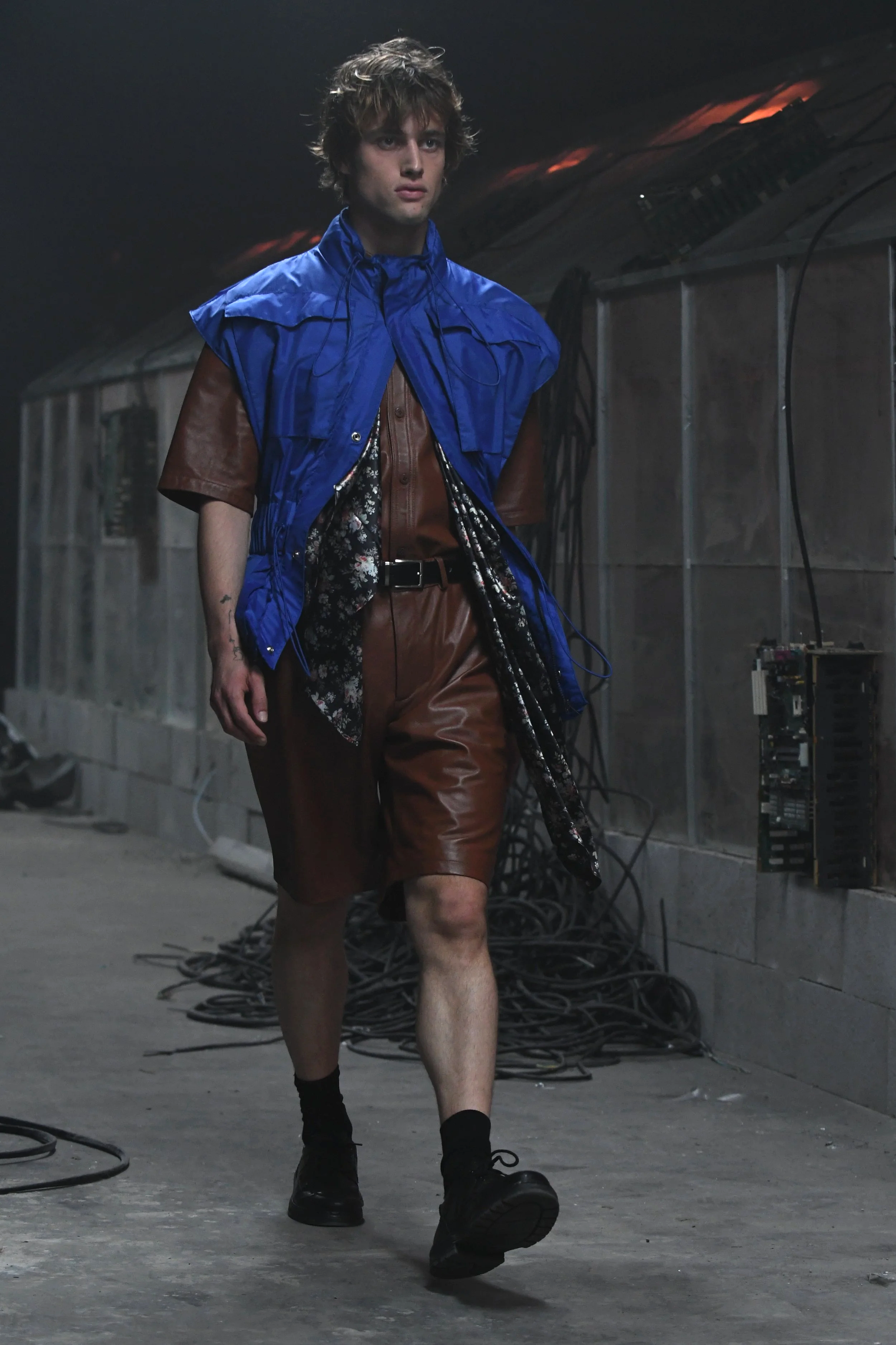 A male model walks down a dimly lit industrial runway wearing a brown leather shirt and matching shorts, with a bright blue sleeveless vest over it, black shoes, and black socks.