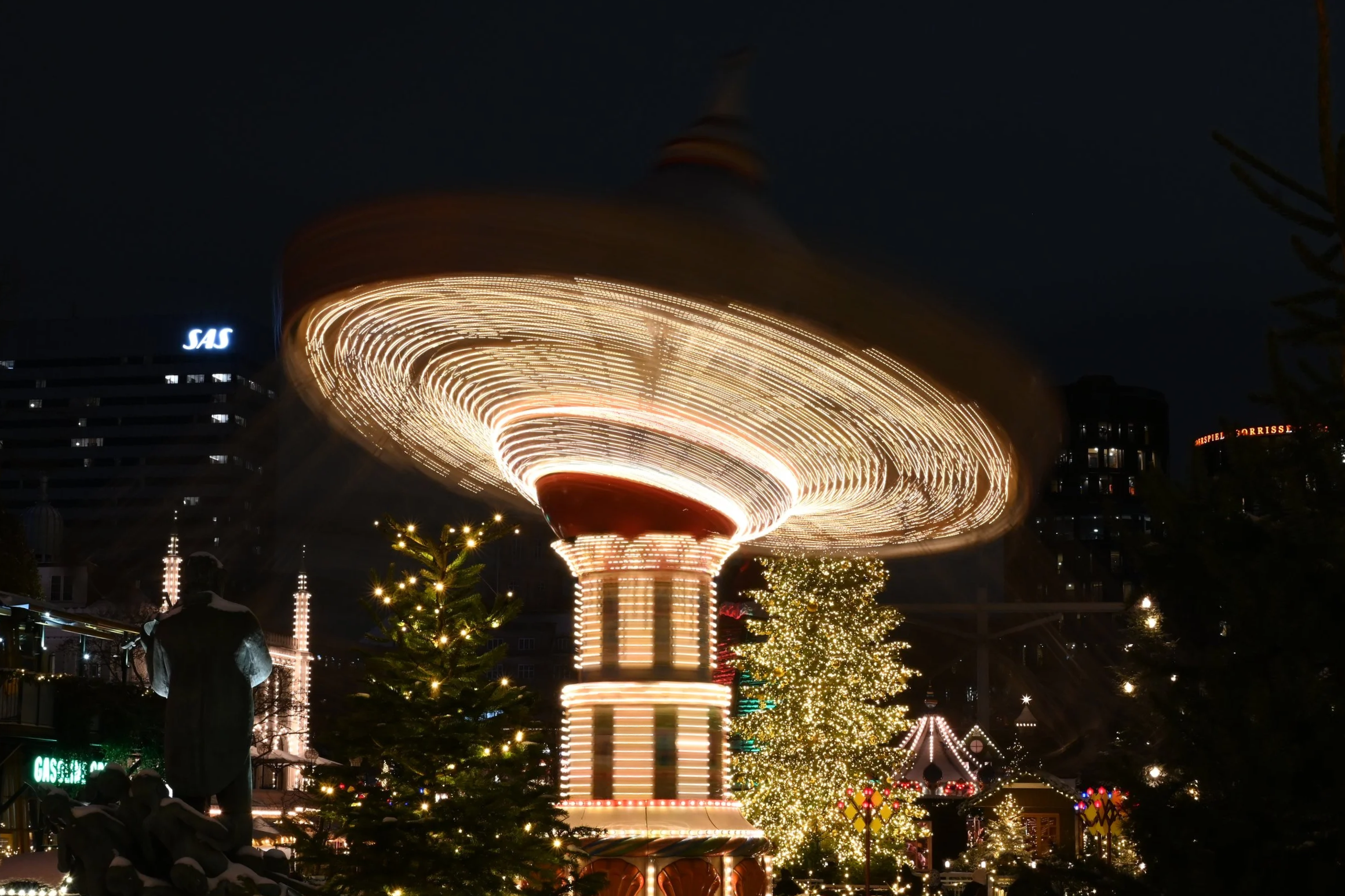 A brightly lit amusement park ride spinning at night with festive holiday lights and Christmas trees around.