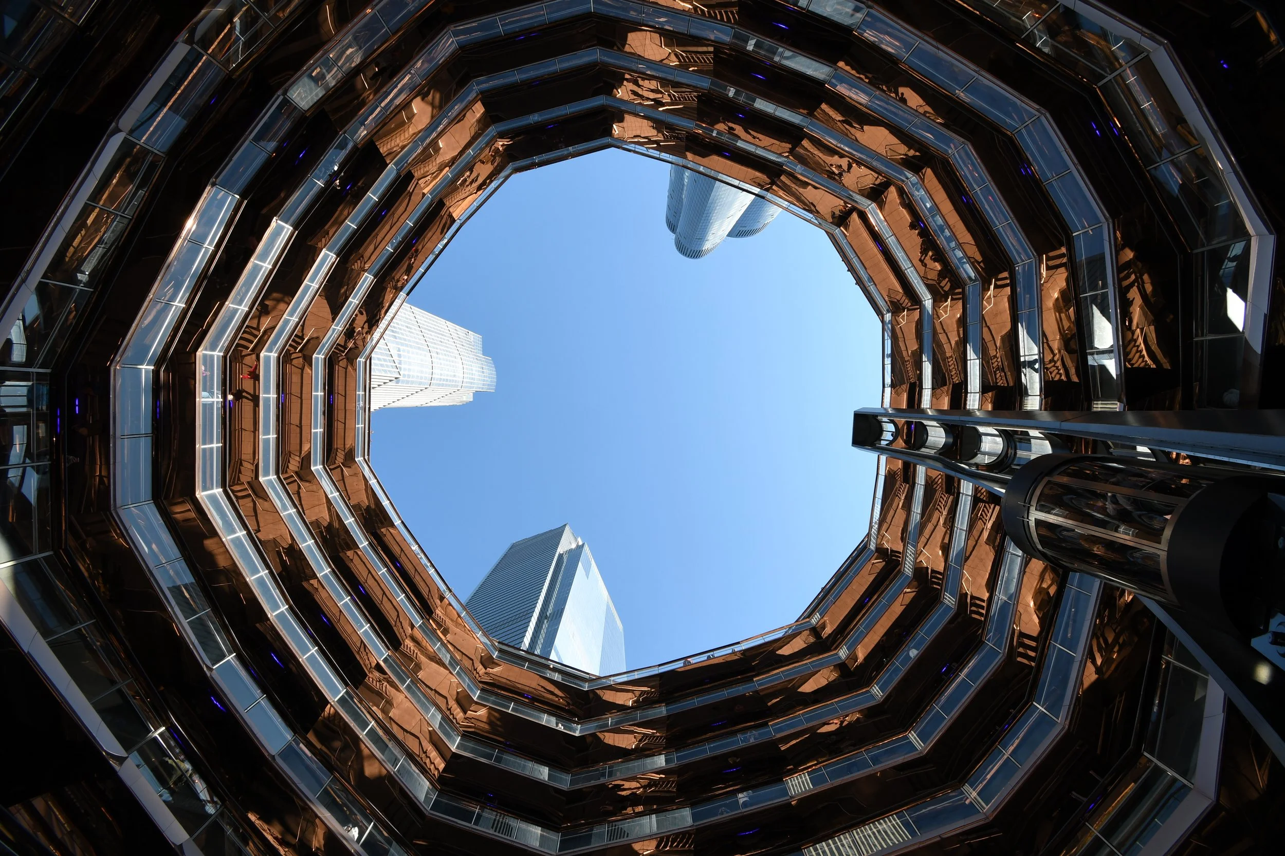 View looking up from the center of a modern building's atrium, showcasing its multi-story glass and metal structure. Skyscrapers are visible through the open top, with a clear blue sky above.