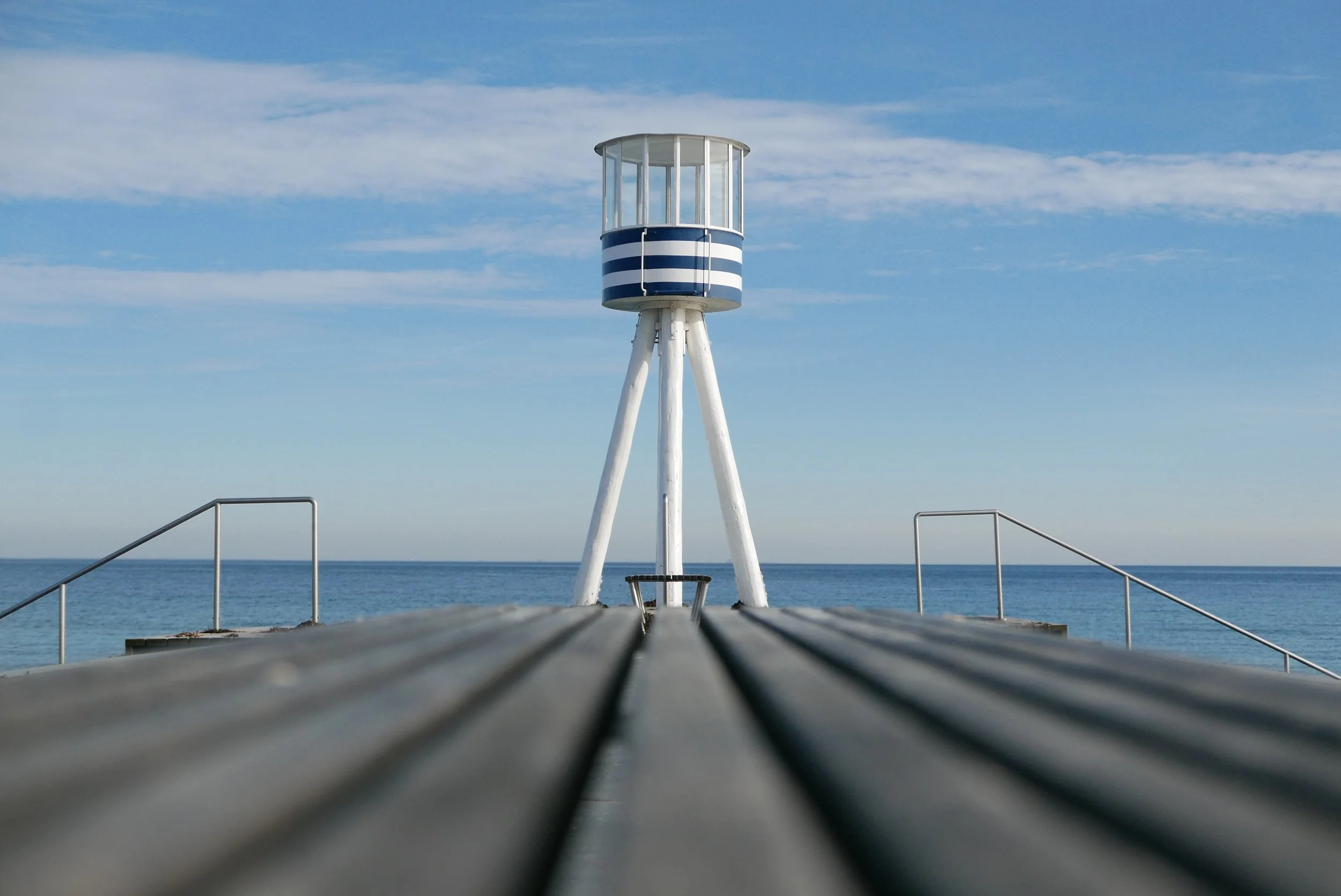A wooden pier leading to a small lighthouse with a blue and white striped exterior, overlooking the ocean under a partly cloudy sky.