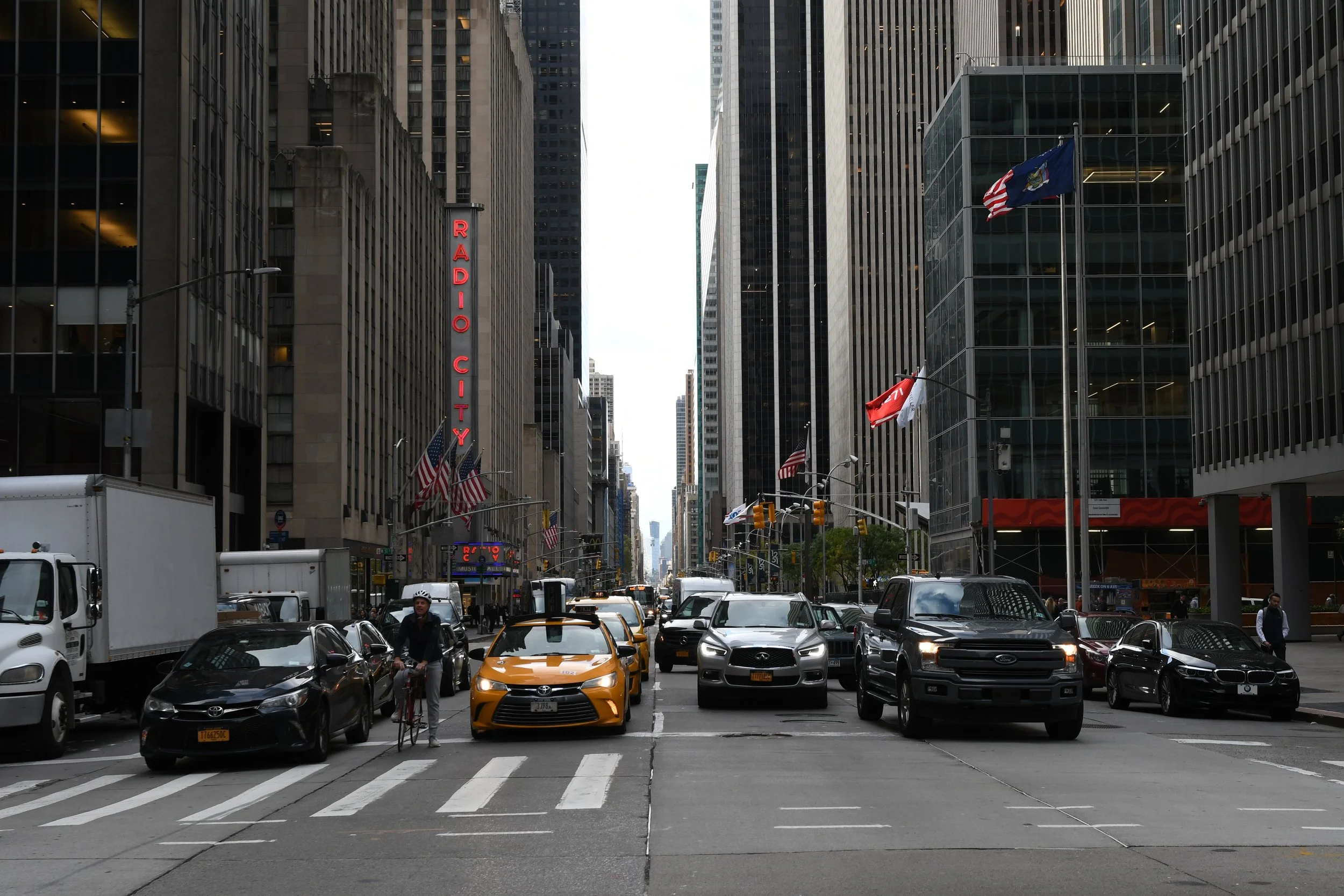 City street scene with tall skyscrapers, a traffic signal, multiple cars including a yellow taxi, a cyclist, with flags including American flags, and a large red neon sign that reads 'Radio City' on a building.