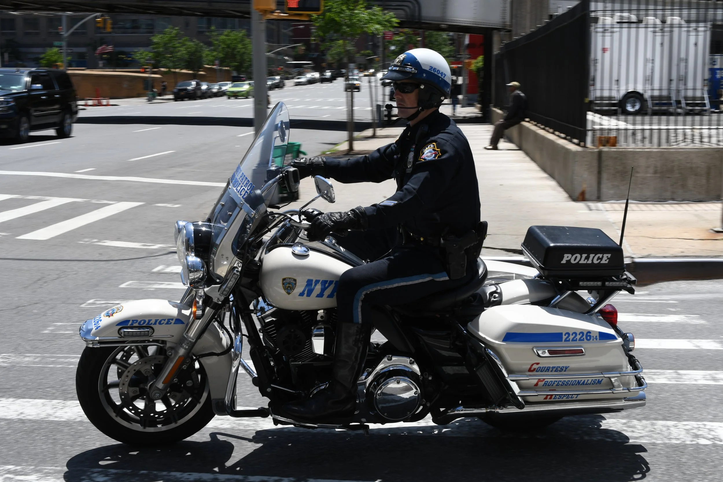 NYC police officer riding a police motorcycle on city street, wearing black uniform and helmet, with trees and pedestrians in background.