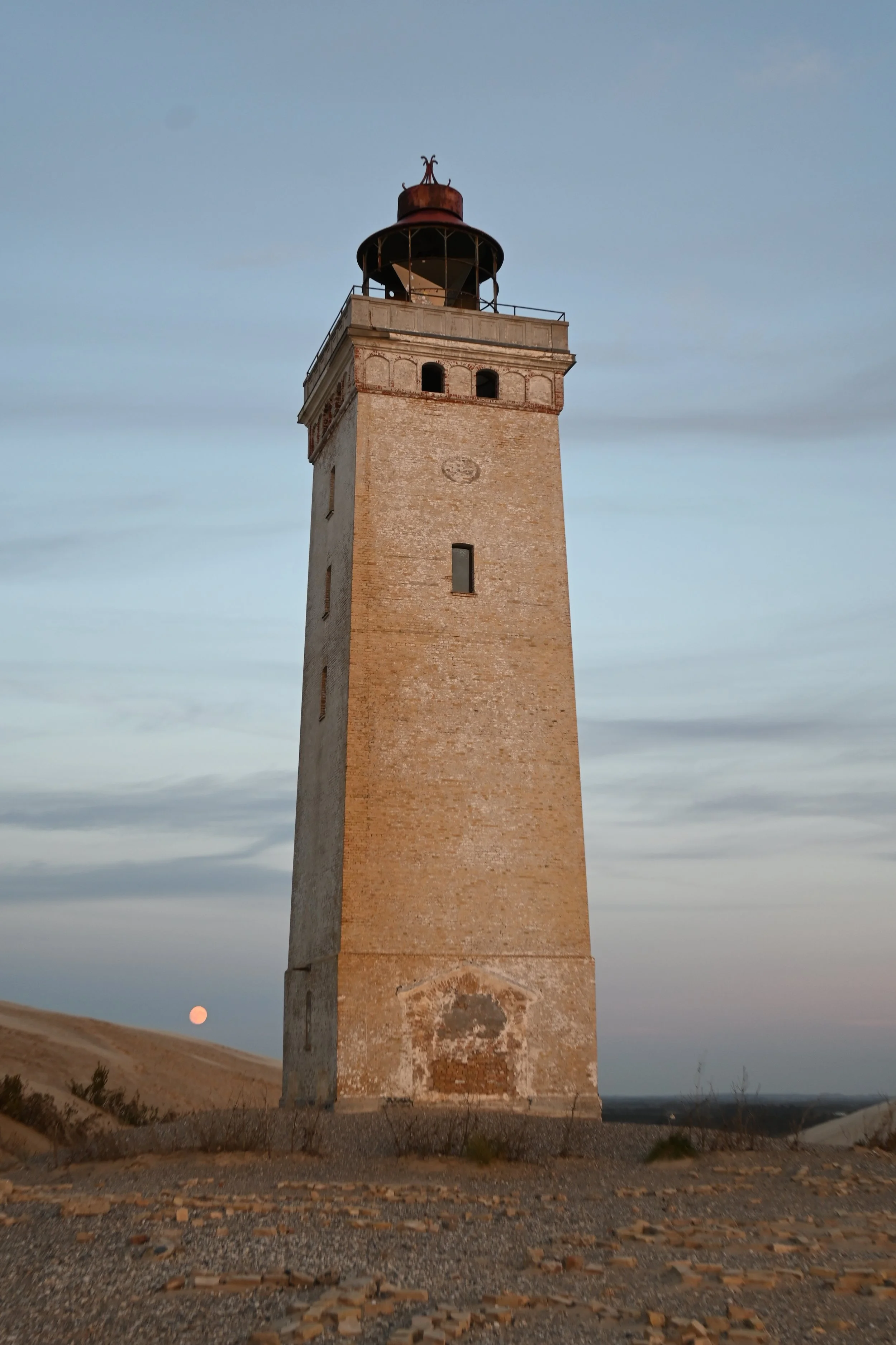 A tall, old brick lighthouse with a rusted metal top, standing on a rocky terrain against a sky with the moon visible in the distant background.