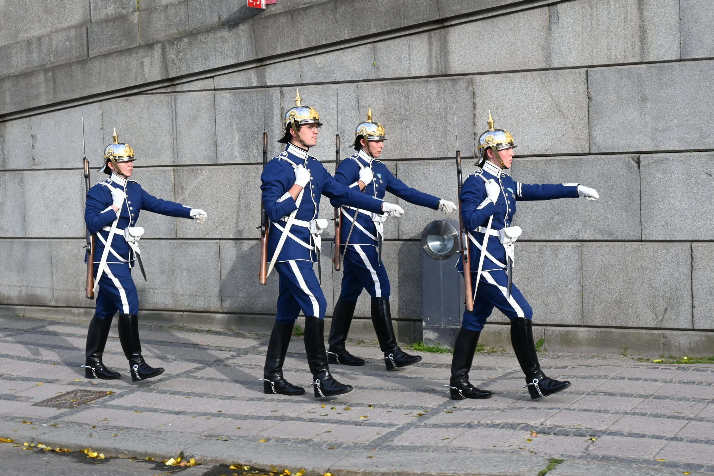 Four uniformed soldiers marching in formation along a city street against a stone wall background.