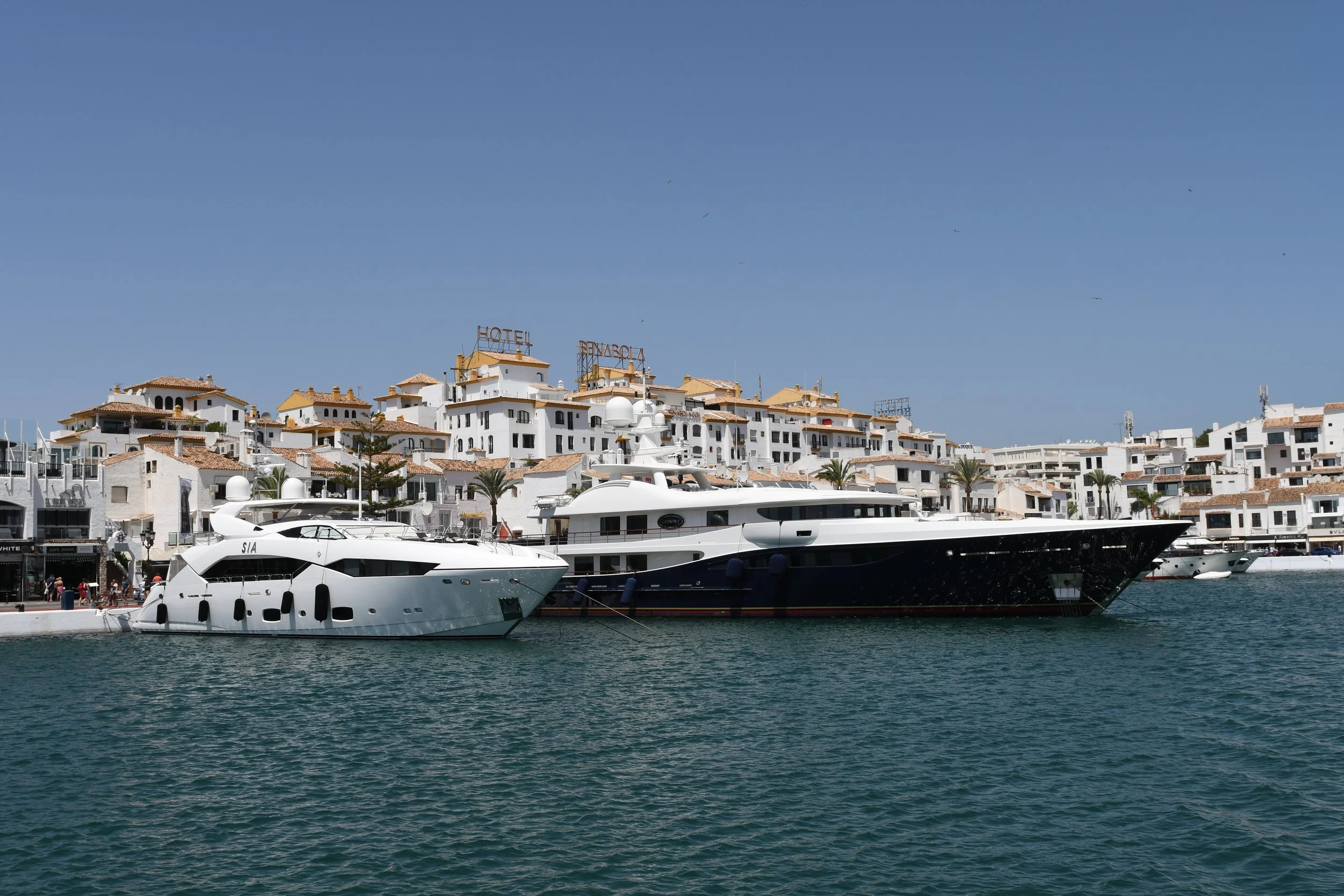 Luxury yachts docked at a marina with white buildings and palm trees in the background, under a clear blue sky.