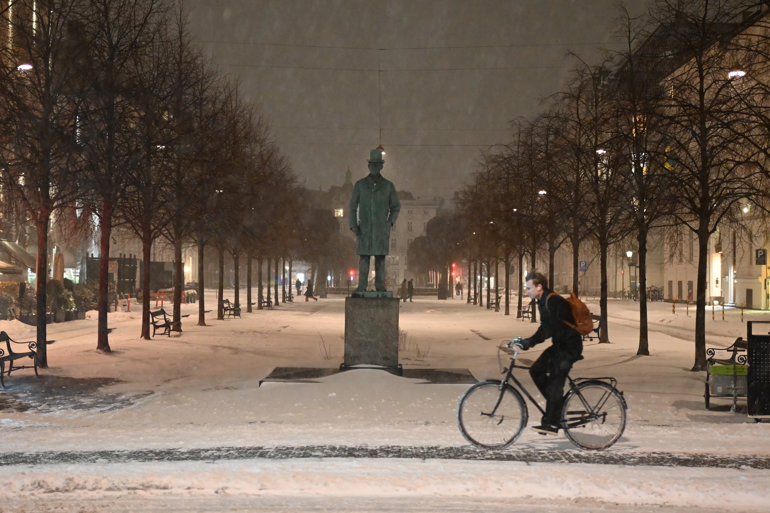 A person riding a bicycle past a statue in a snow-covered park at night, with leafless trees and streetlights illuminating the scene.