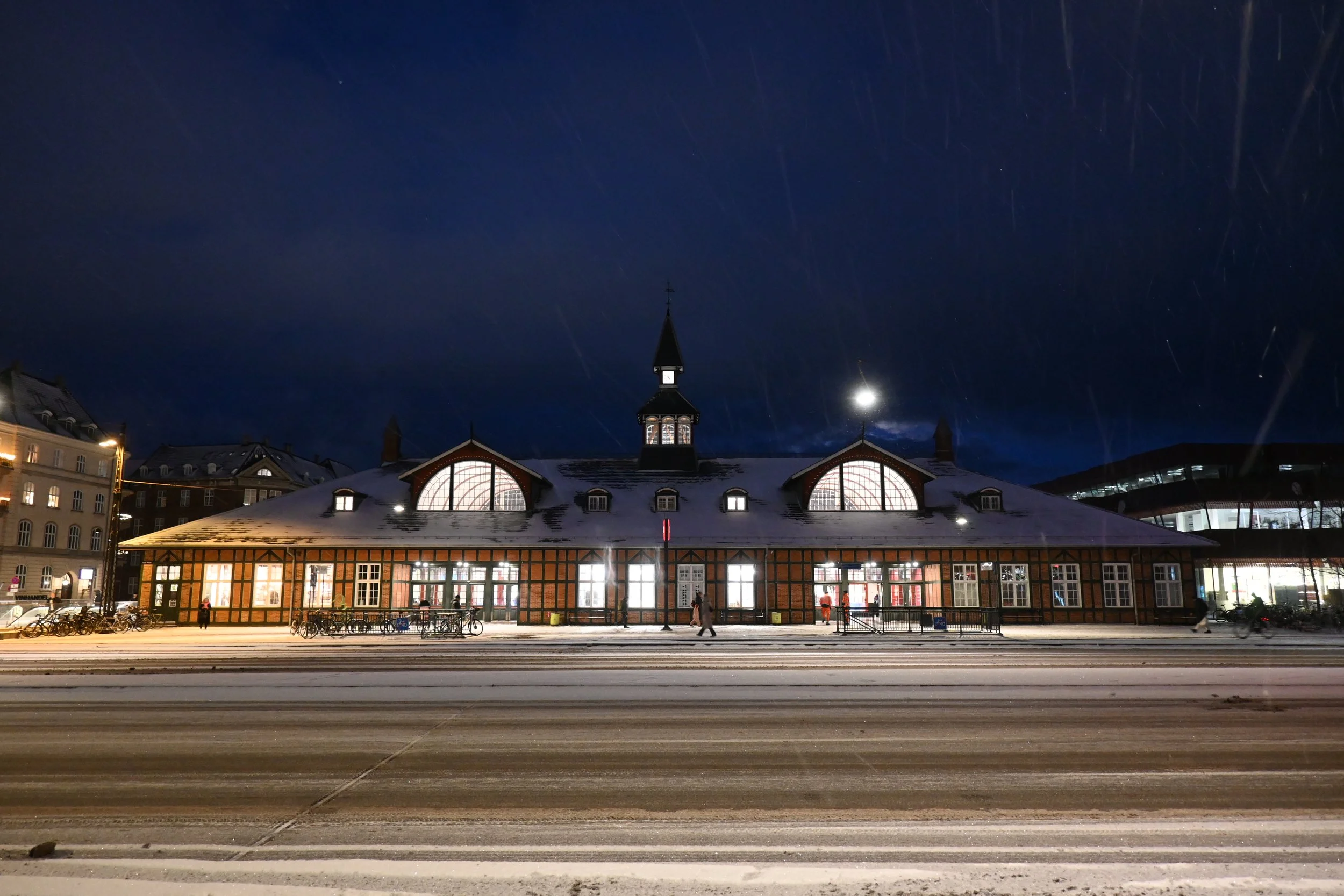 Night view of a historic train station building with lit windows, snow on the roof, and a dark sky with star trails.