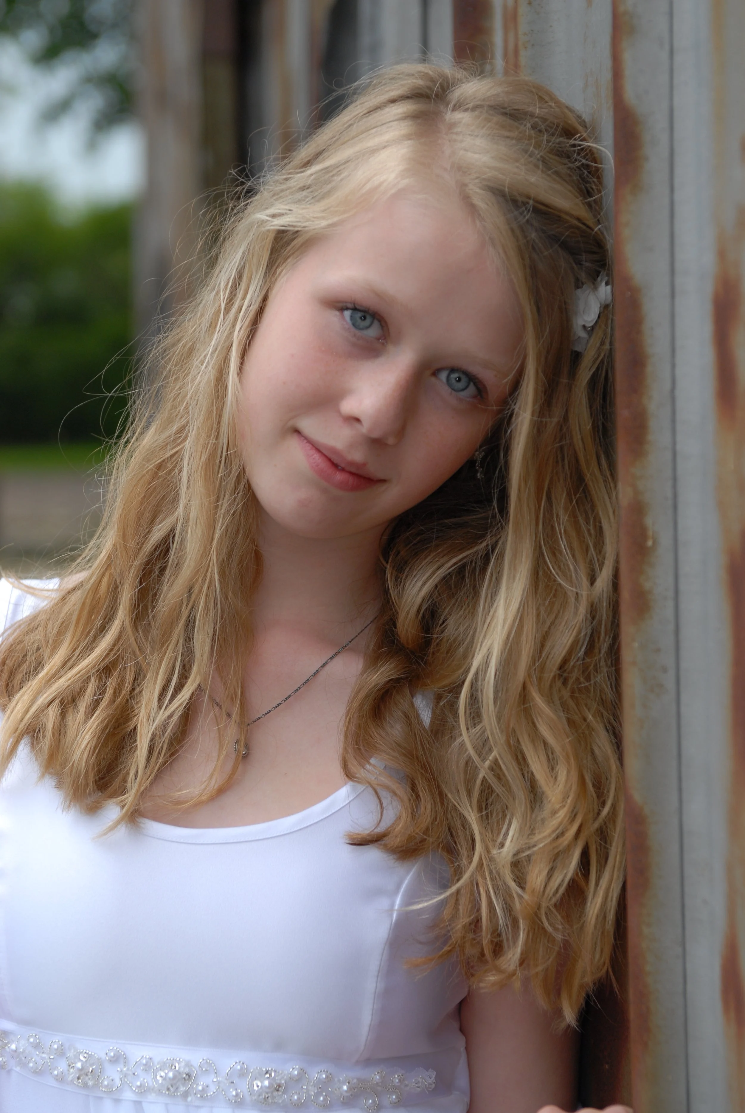 A young woman with long blonde hair and blue eyes leaning against a rusted metal wall outdoors.