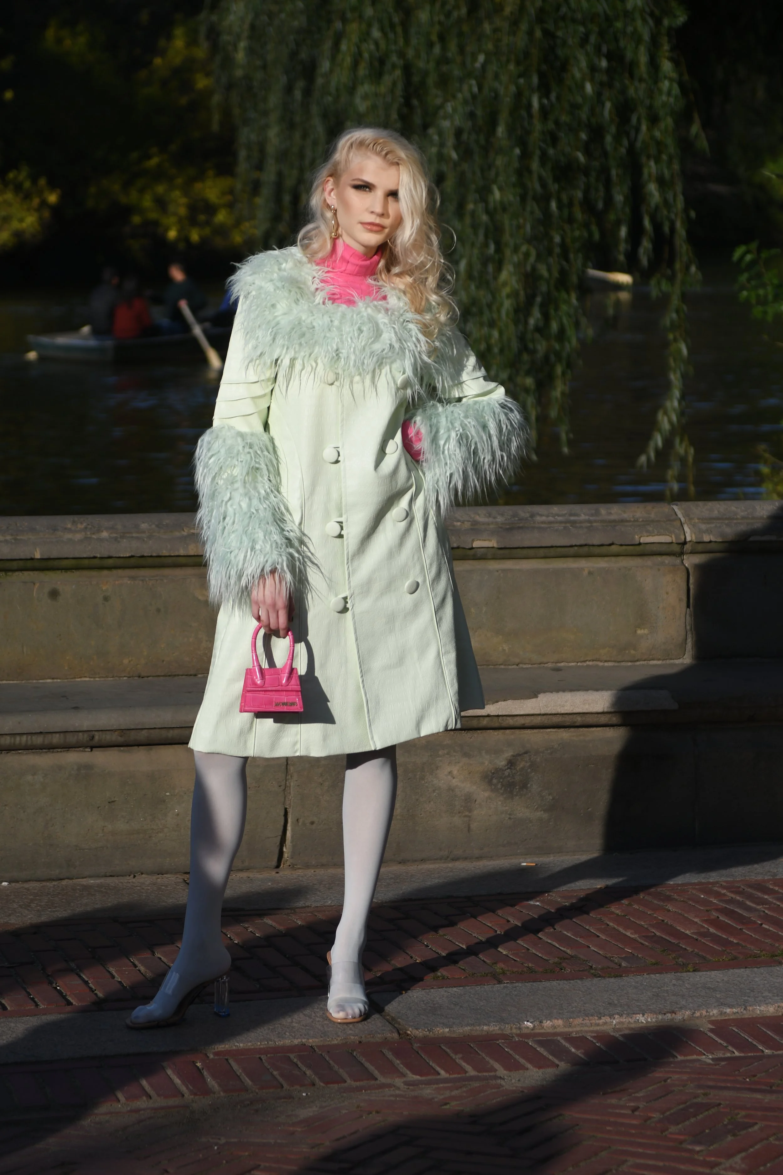 A woman standing outdoors by a stone ledge, wearing a light-colored coat with fluffy fur accents, white tights, and clear high heels. She holds a small pink handbag and has long, wavy blonde hair. There is water and a small boat with people in the ba