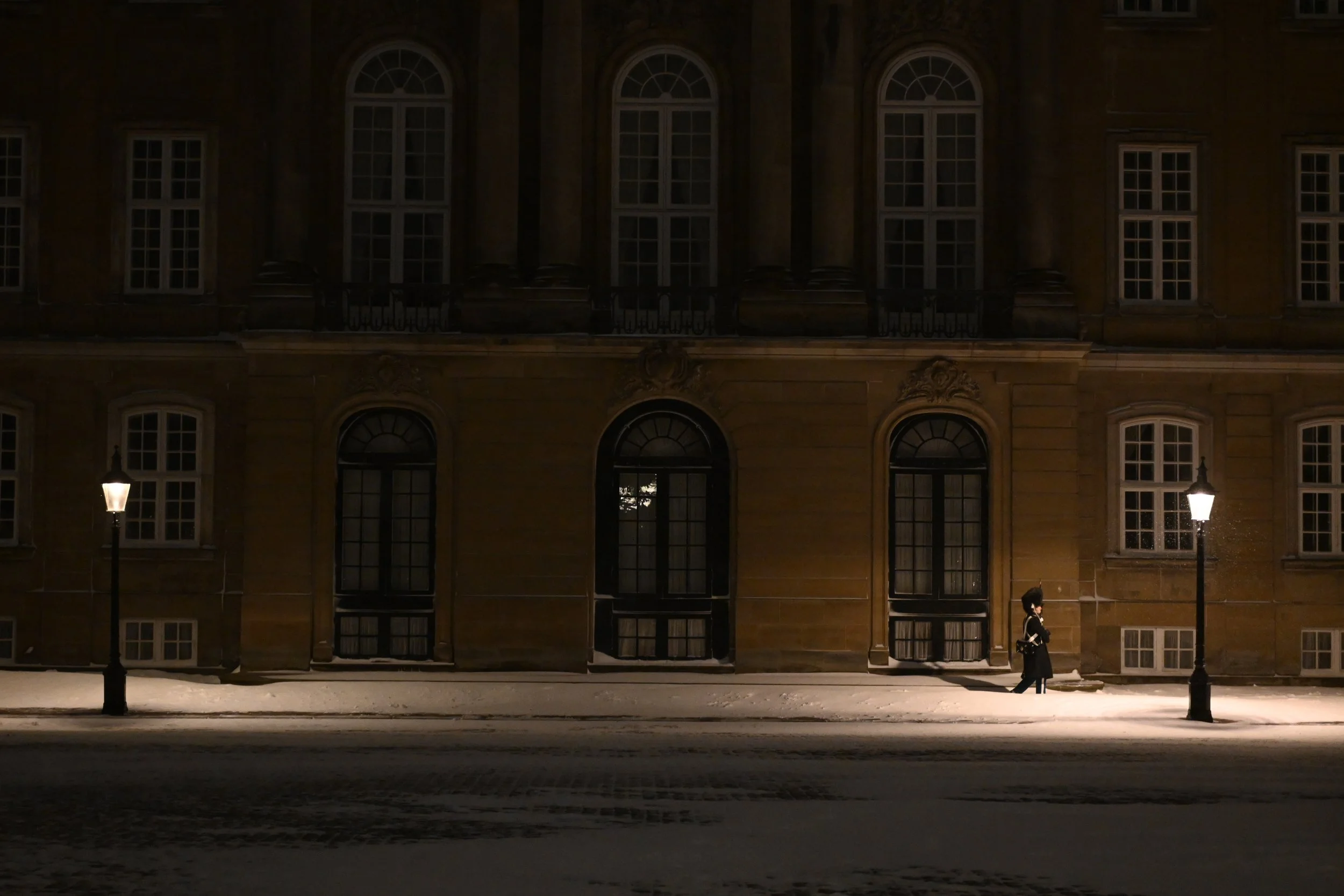 A person walking on a snowy sidewalk at night, between two lit street lamps, in front of a large, historic stone building with numerous windows.