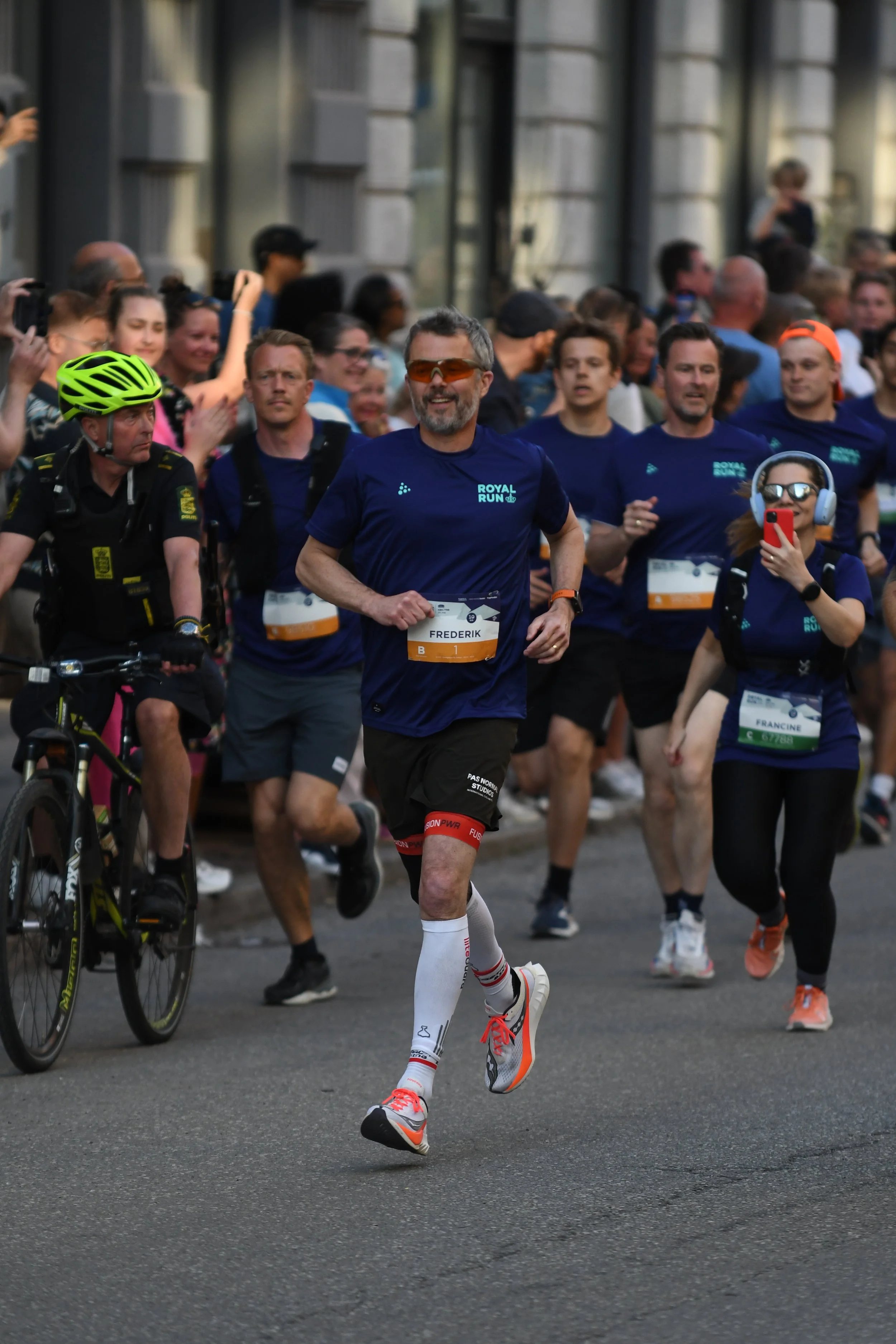 A group of runners participating in a marathon, led by a man with gray hair and a beard wearing a blue shirt, black shorts, compression socks, and running shoes, along with other runners and spectators on the street.