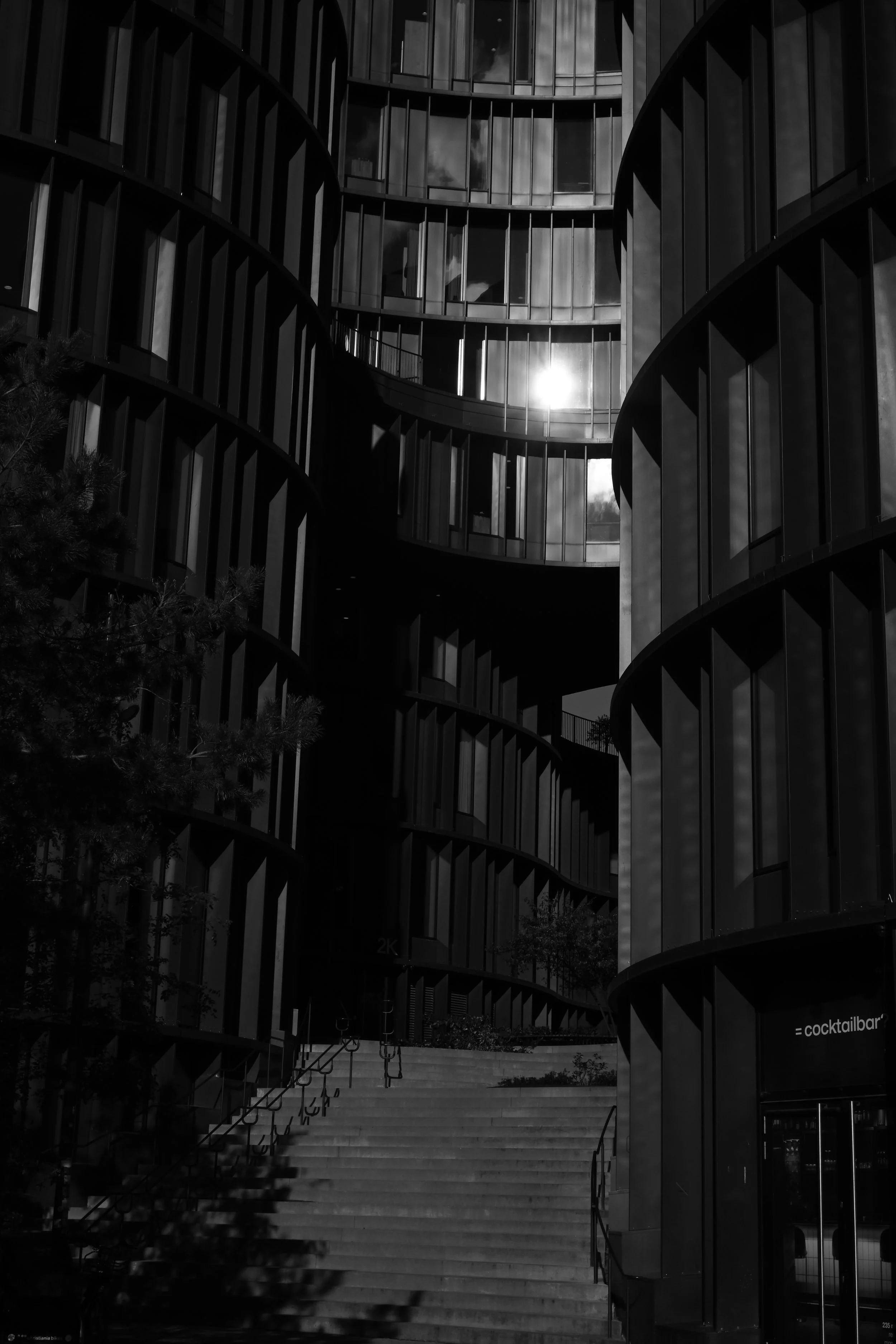 Black and white photo of modern multi-story building with curved glass walls and a staircase leading up to the entrance.