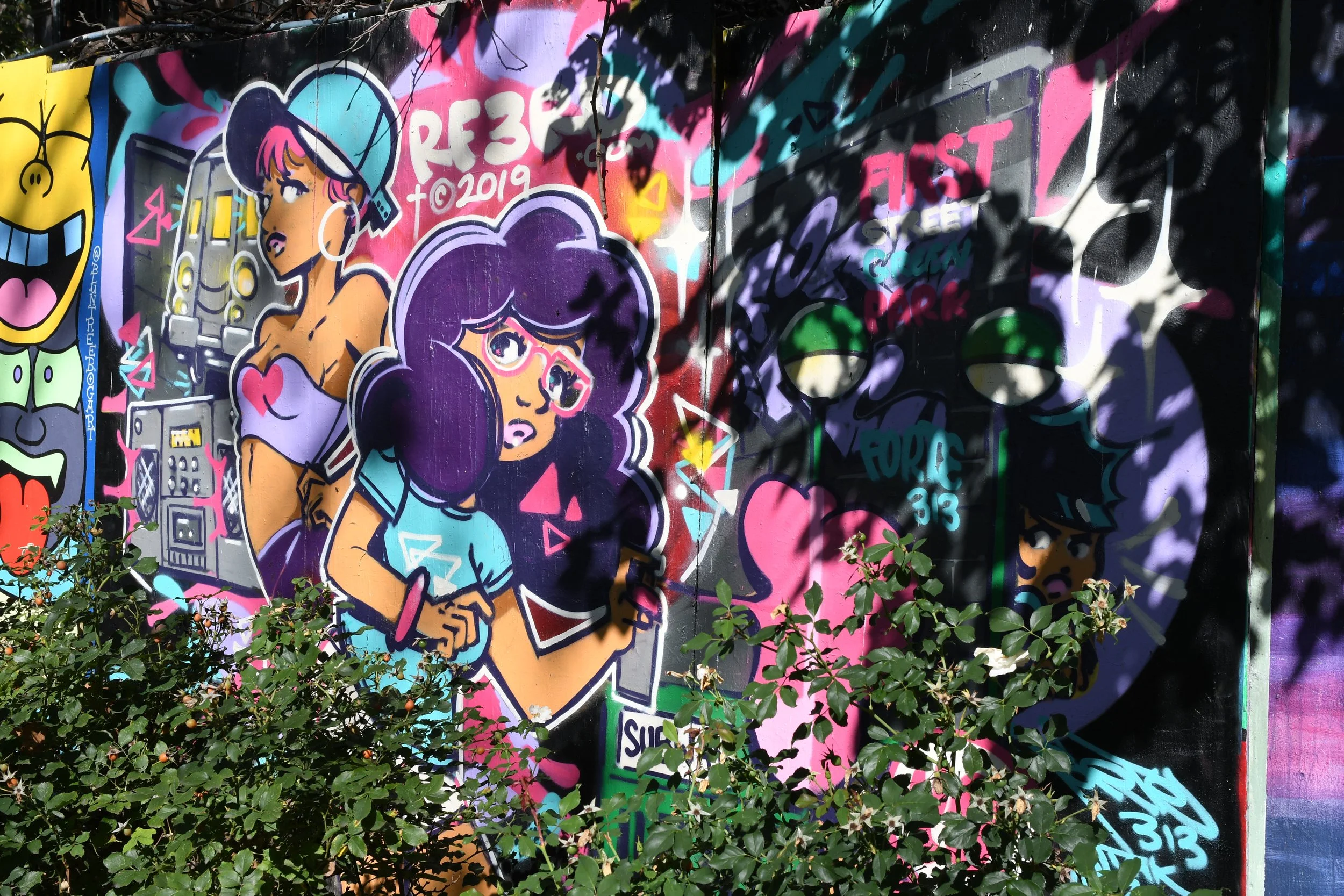 Colorful graffiti mural featuring cartoon-style characters, including a girl with purple glasses and large hair, another girl with a heart-shaped pendant, and a character with spiky hair and sunglasses, surrounded by abstract shapes and text.