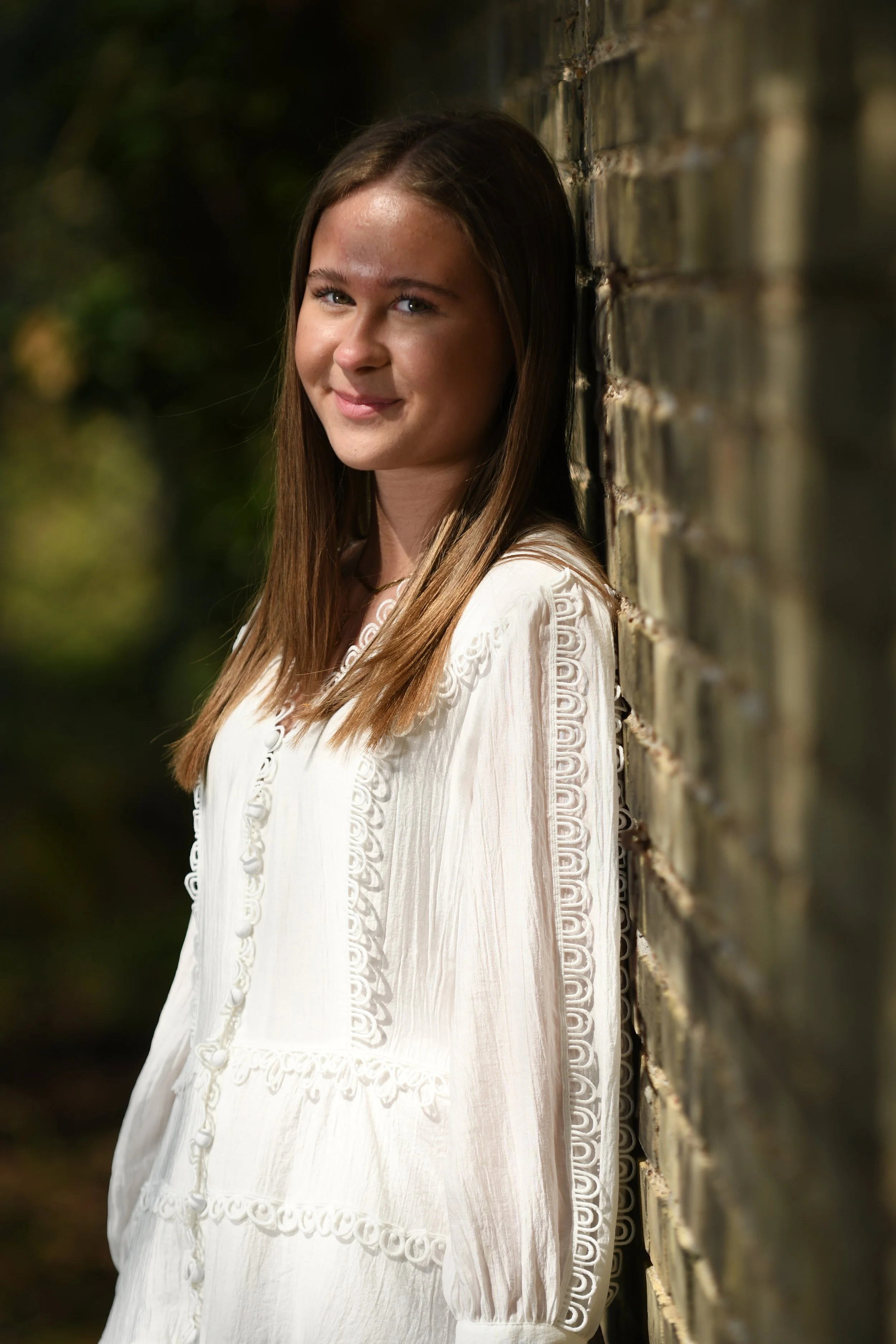 A young woman leaning against a brick wall outdoors, smiling at the camera. She has long brown hair and is wearing a white embroidered blouse.