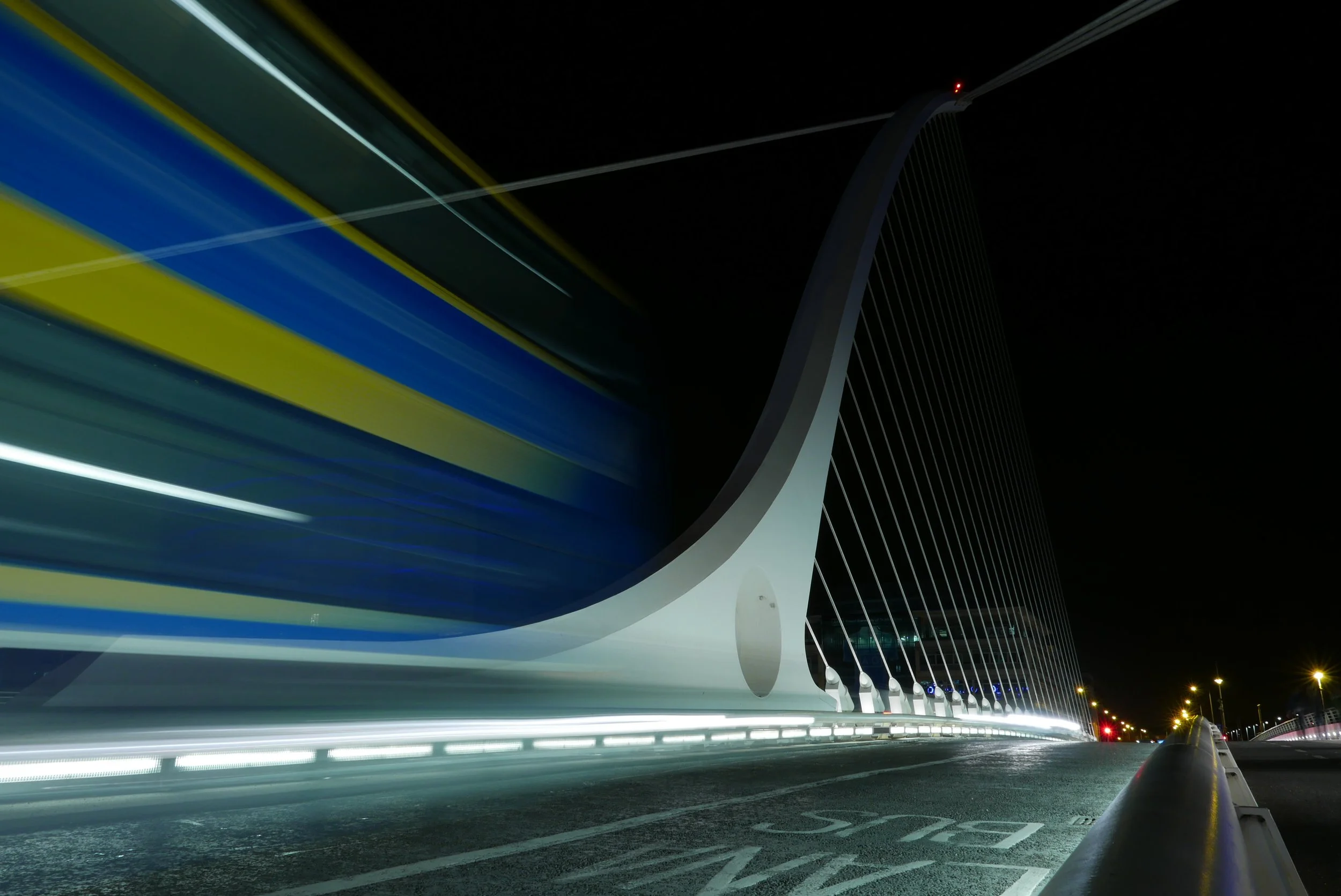 Night view of a modern cable-stayed bridge with illuminated struts and cables, captured with light trails from passing vehicles.