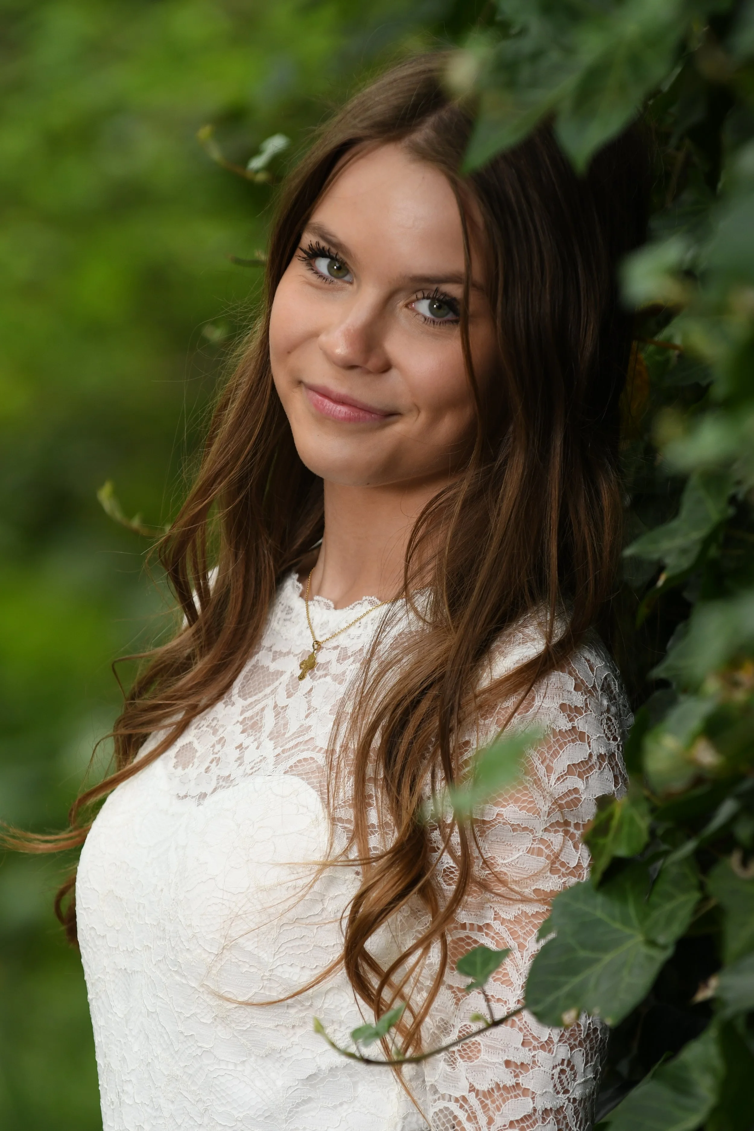 A young woman with long brown hair and green eyes, wearing a white lace dress, smiling softly and leaning against green leafy foliage outdoors.