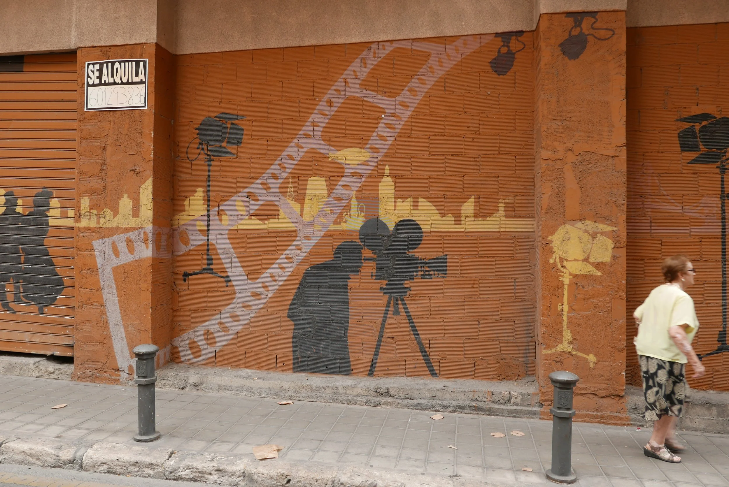 Street art mural of a person filming with a vintage movie camera, silhouette style, on a brick wall. There are additional silhouettes and a skyline in the background, with construction lights and a 'Se Alquila' sign in the upper left corner.